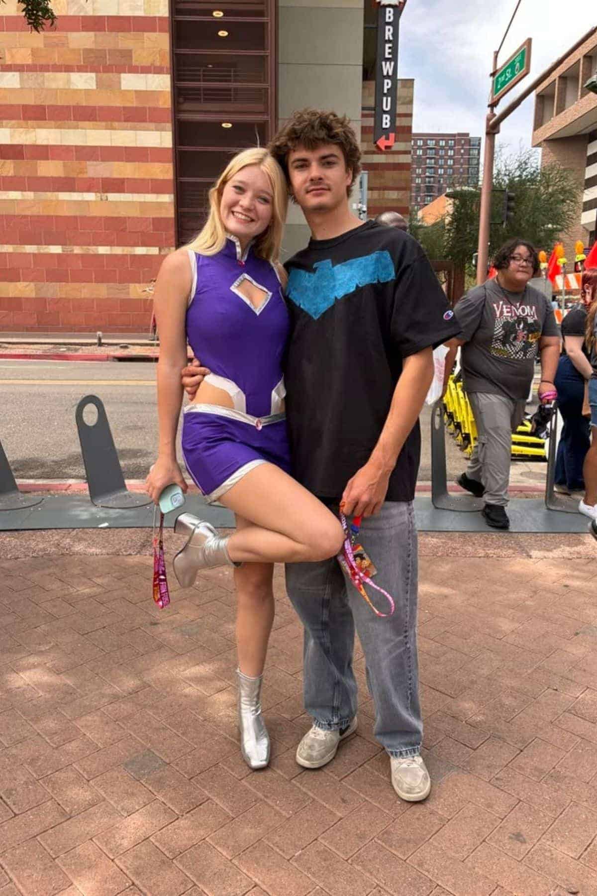 Two people pose on a city sidewalk; one wears a purple costume and silver boots, the other wears jeans and a black shirt with a blue bird symbol.