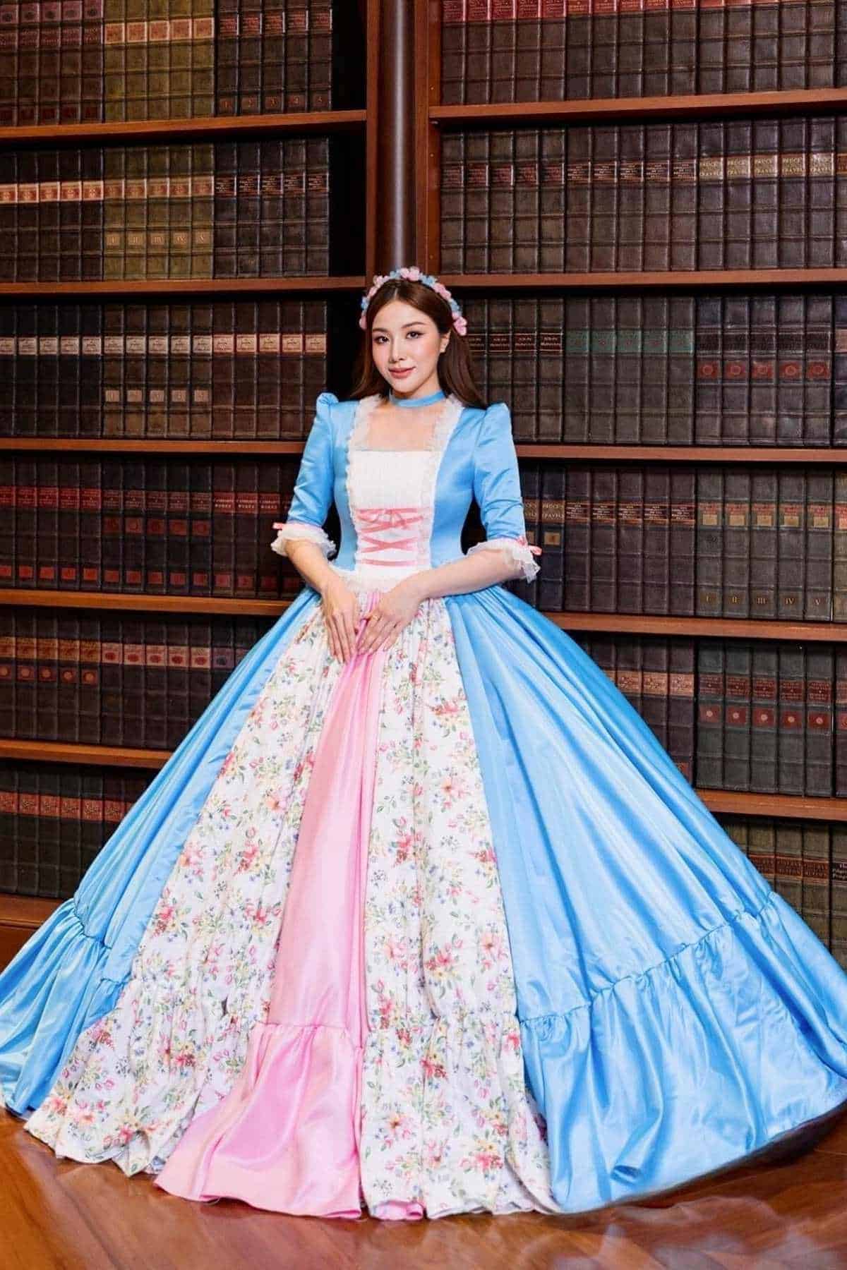 A woman in a blue, pink, and floral ball gown stands in front of tall bookshelves filled with books.