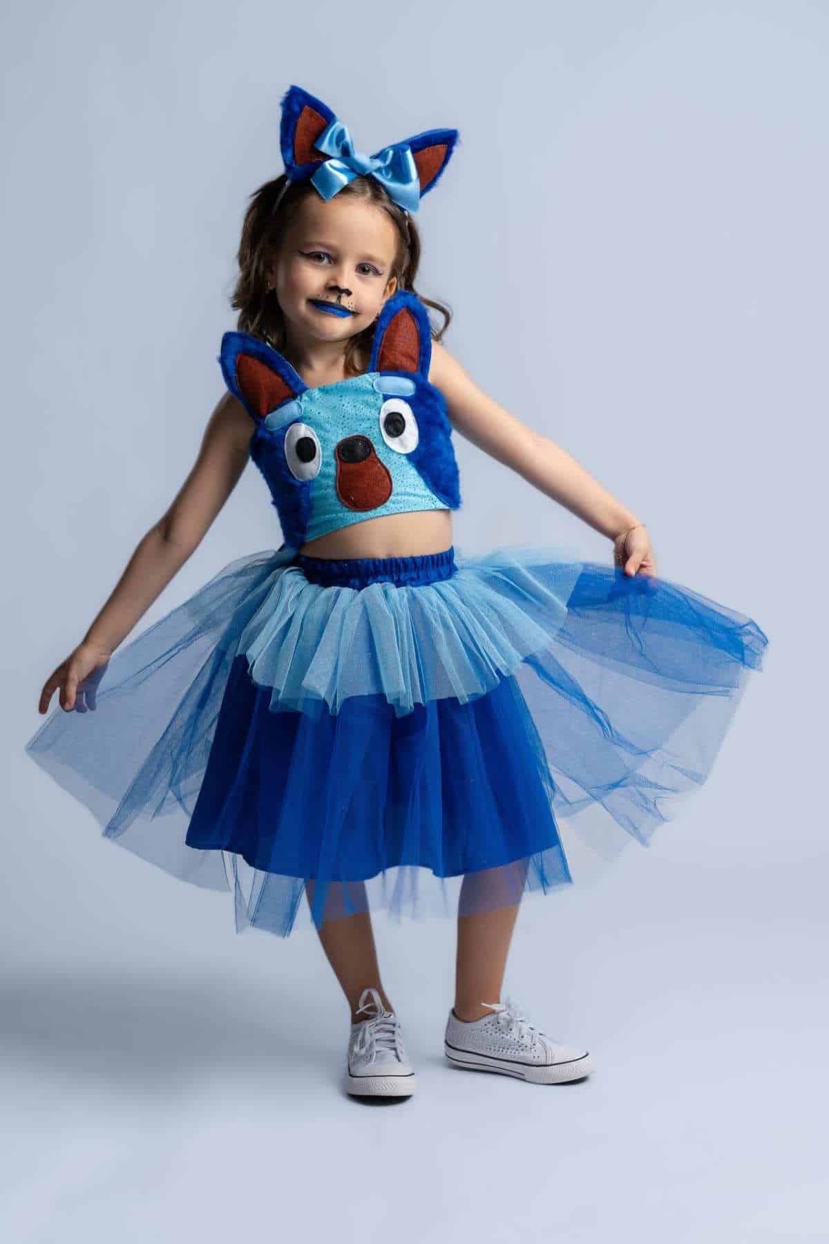 Child in a blue tutu and animal-themed top with matching ears headband, posing against a plain background.