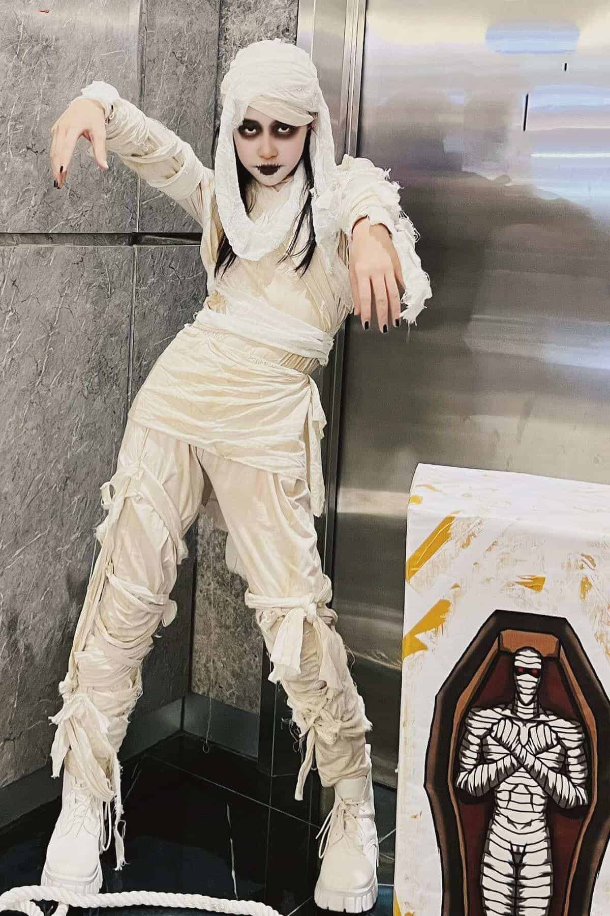 Person dressed as a mummy with white bandages, dark eye makeup, and pale face poses near a drawing of a mummy in a sarcophagus.