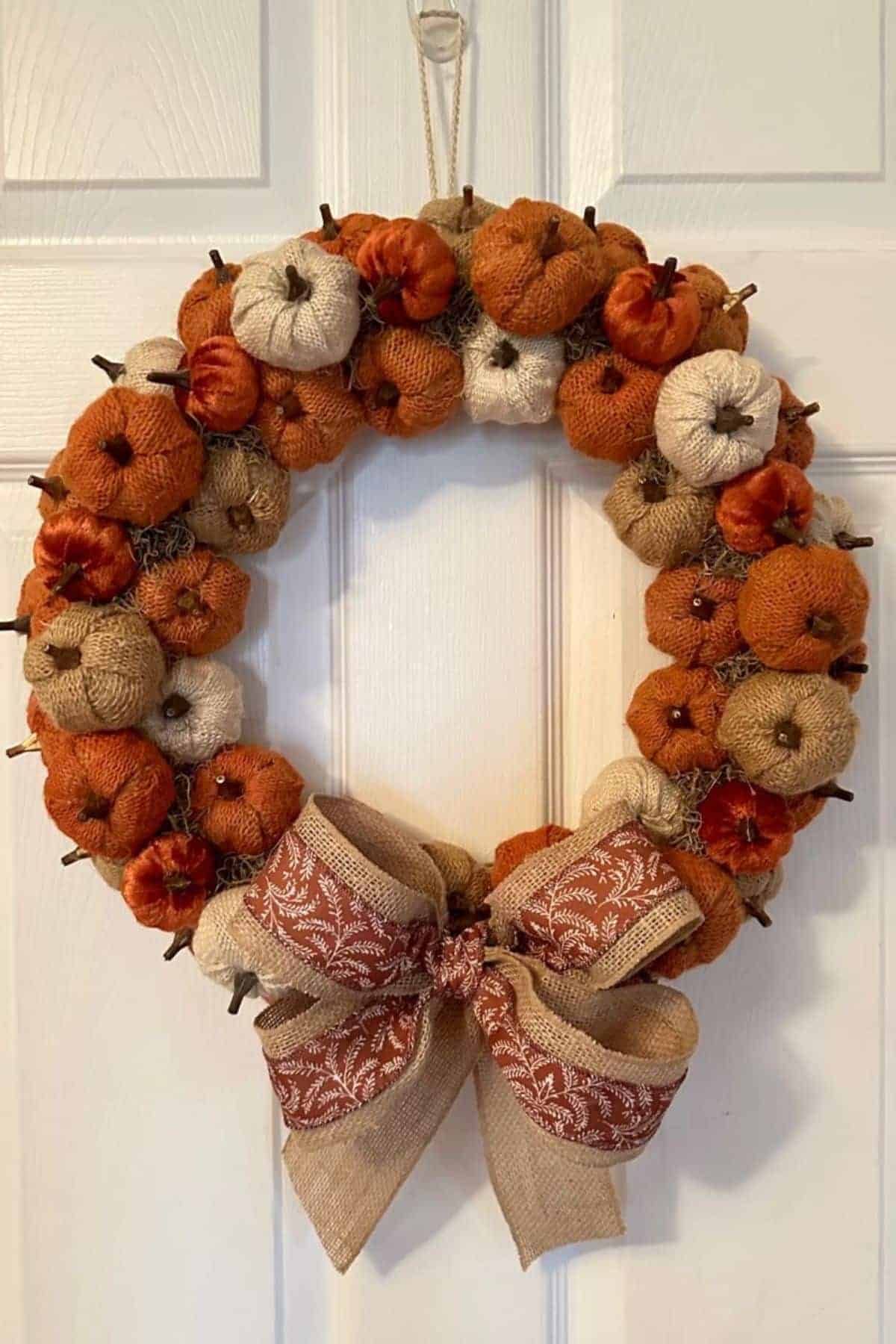 A wreath made of small orange and beige fabric pumpkins with a large burlap bow hangs on a white door.