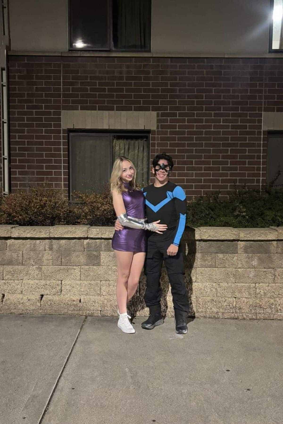 Two people in superhero costumes stand together on a sidewalk at night in front of a brick building, smiling at the camera.