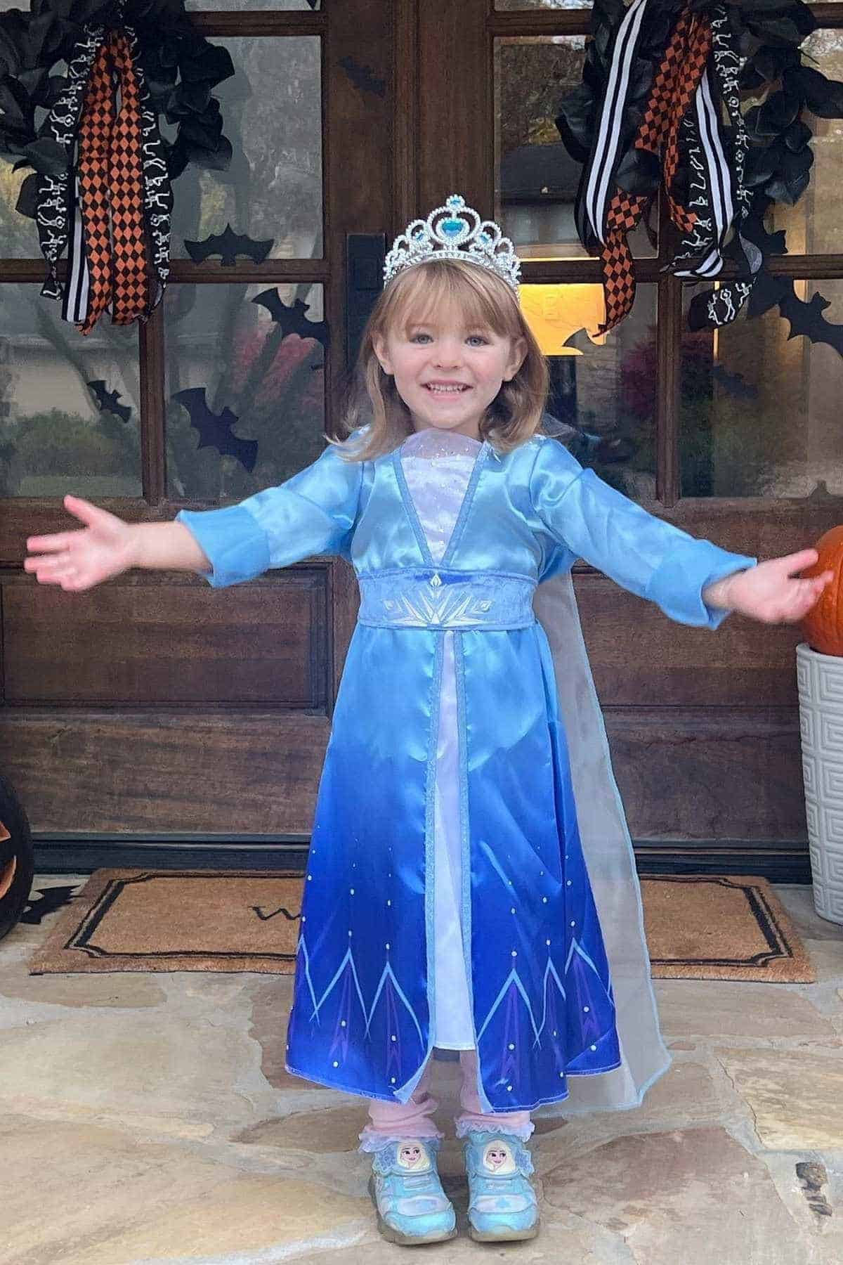 Young child in a blue princess dress and tiara stands with arms open in front of a decorated door with Halloween ribbons and a pumpkin.