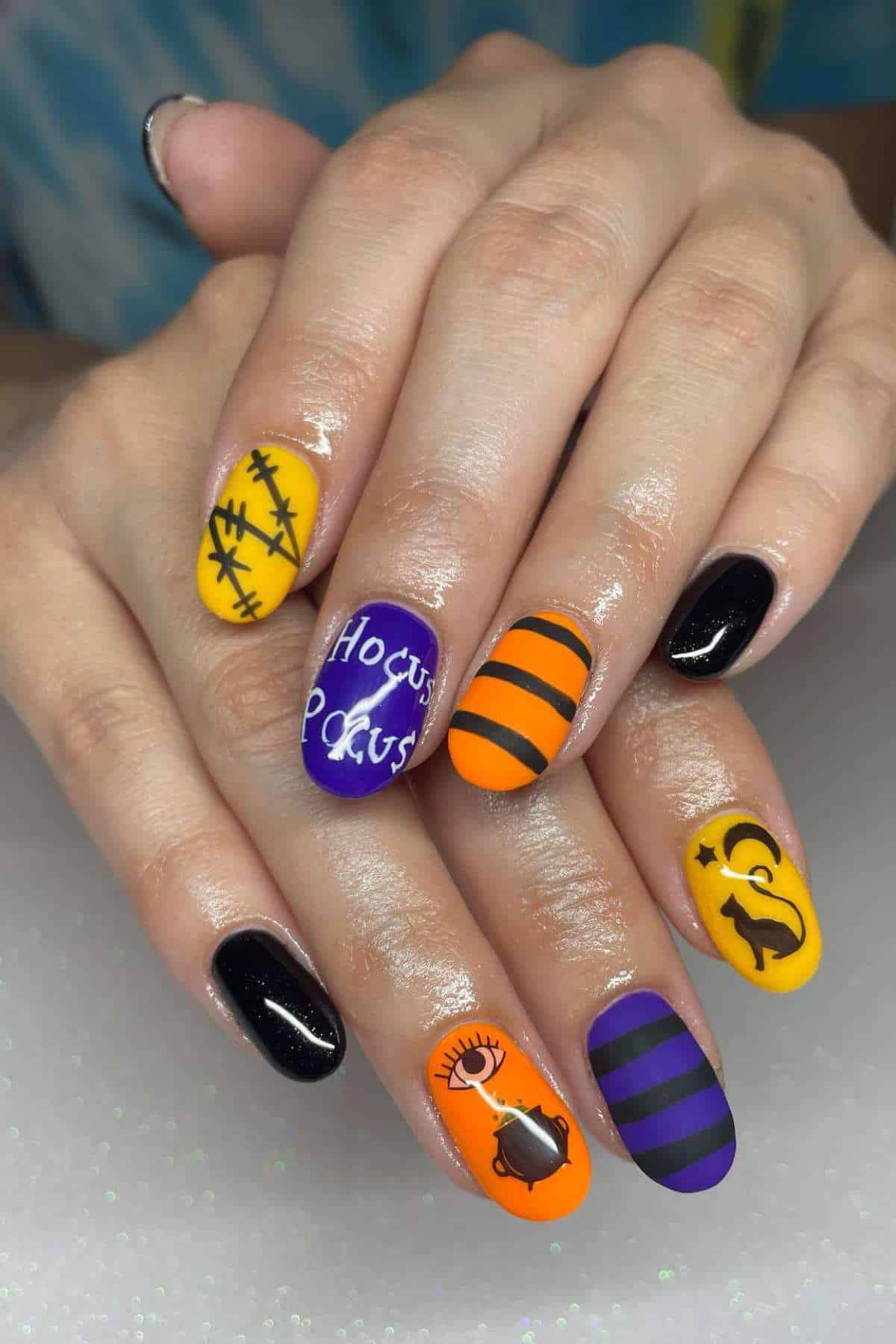 Hands with Halloween-themed nail art, featuring black, purple, and orange colors, stripes, and designs like a cat, moon, barbed wire, “Hocus Pocus,” and an eye.