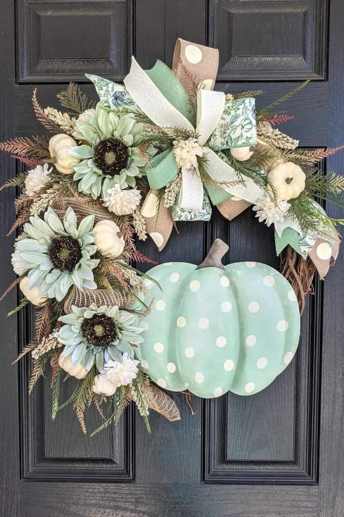 Wreath with white flowers, greenery, decorative ribbons, and a large, light green polka-dotted pumpkin hangs on a black door.