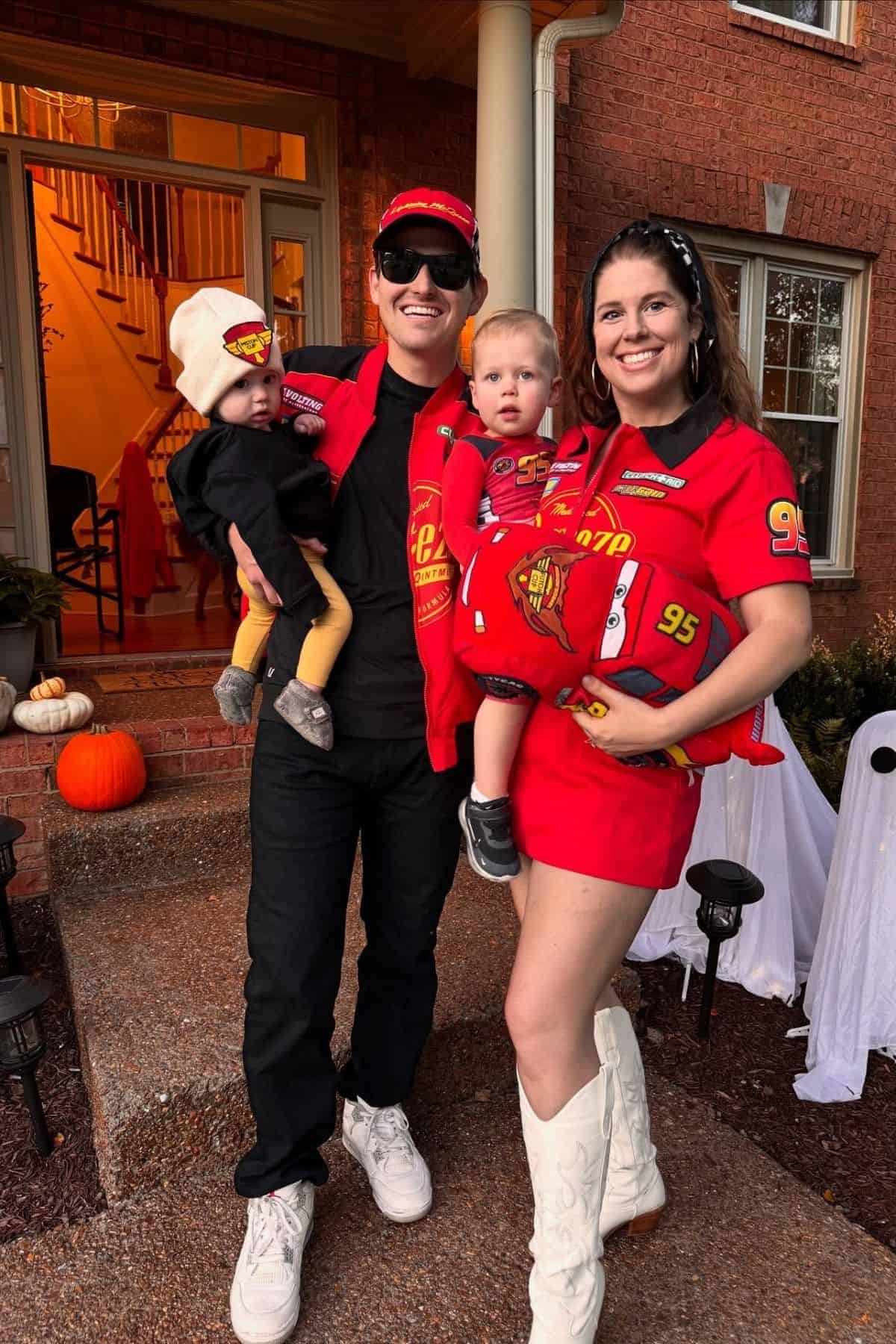 A family of four in matching red racing-themed outfits stands outside a brick house decorated for Halloween with pumpkins and ghost decorations.