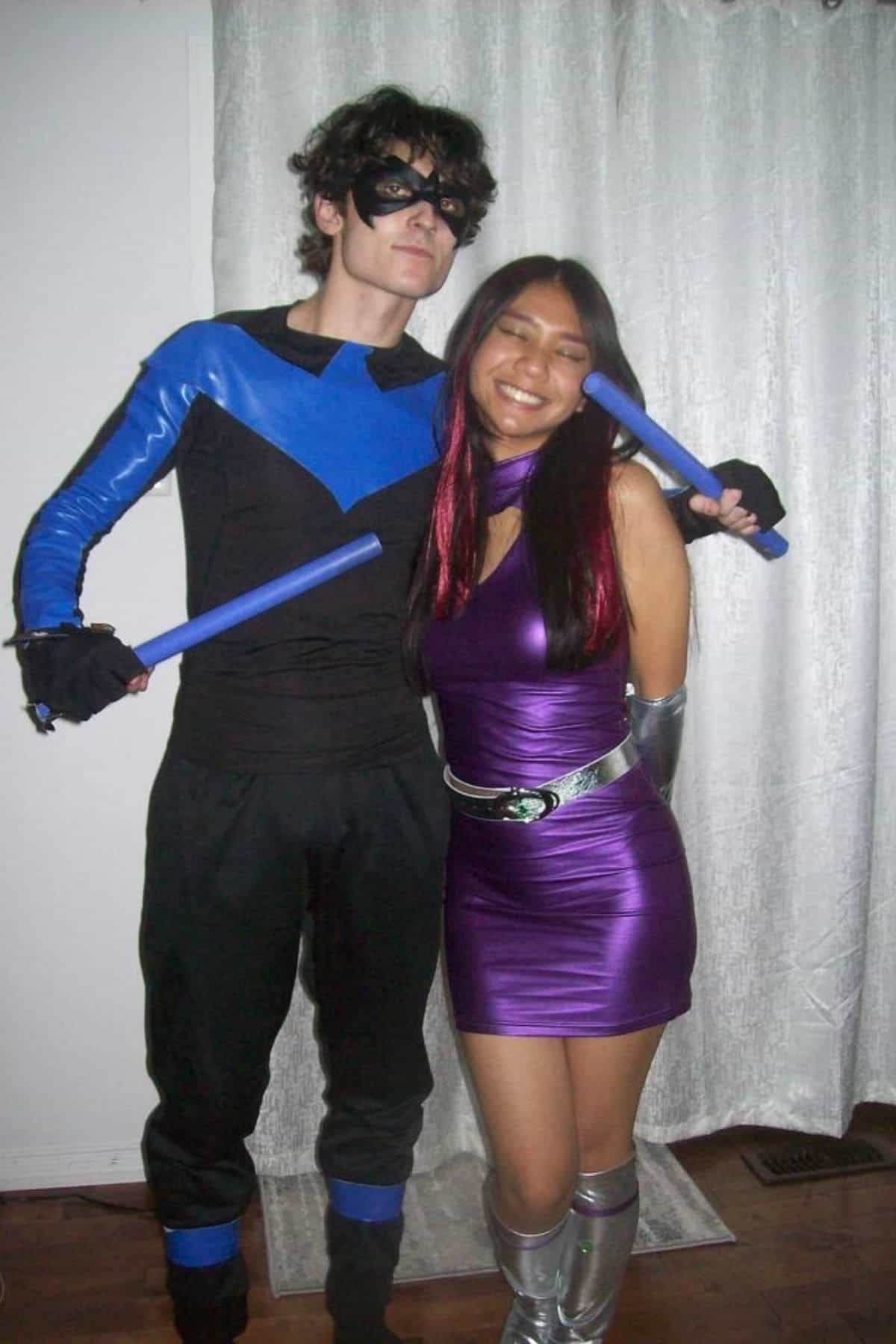 Two people in superhero costumes pose indoors; one wears a black and blue outfit with a mask and batons, the other wears a shiny purple dress with silver accessories and boots.