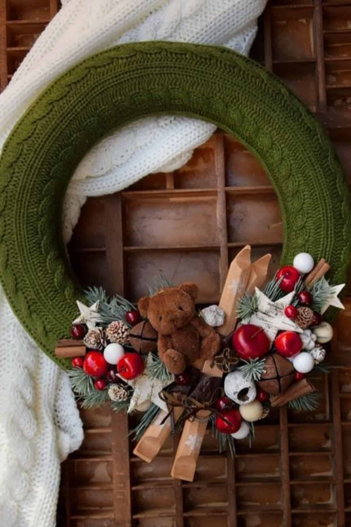 Green knit wreath decorated with a small brown teddy bear, skis, pine branches, red and white ornaments, and pinecones, hanging on a wooden surface with a white knit scarf.