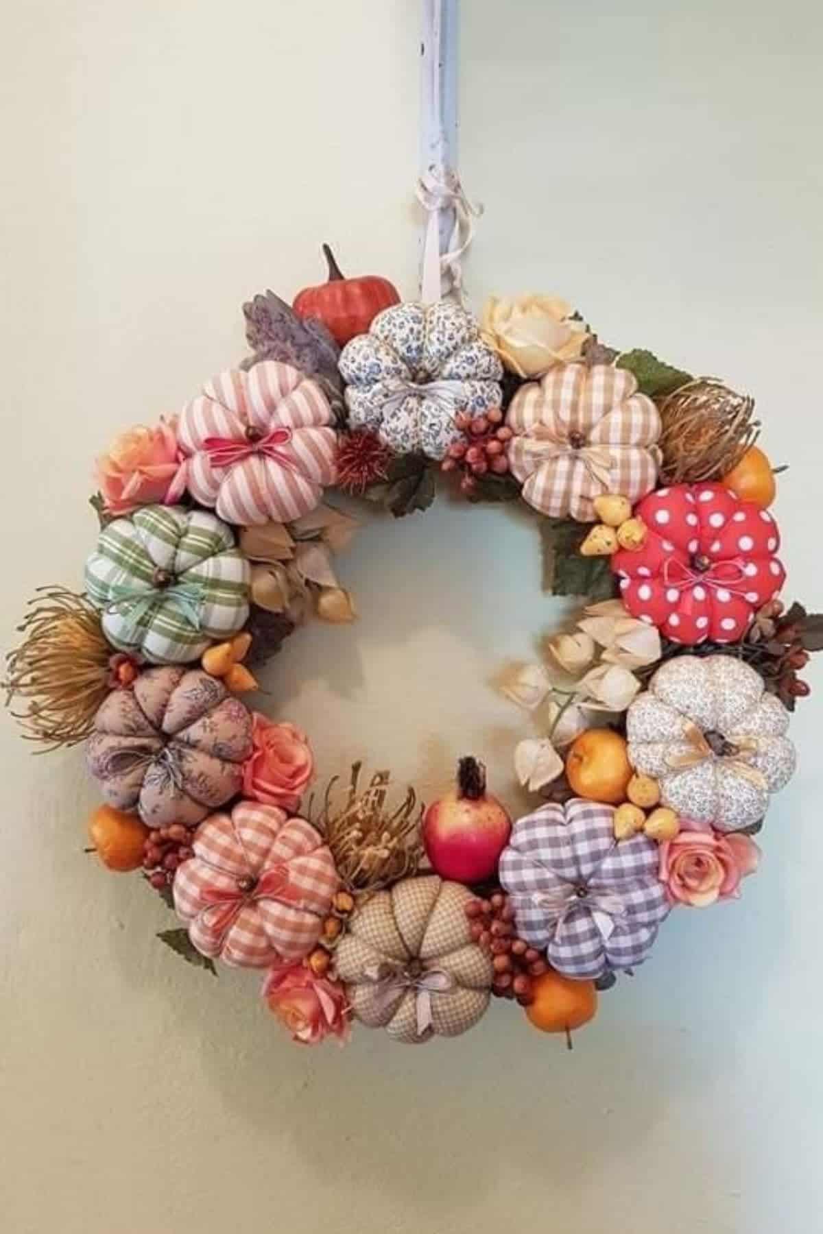 Wreath decorated with fabric pumpkins, artificial fruits, flowers, and leaves, hanging on a light-colored wall.