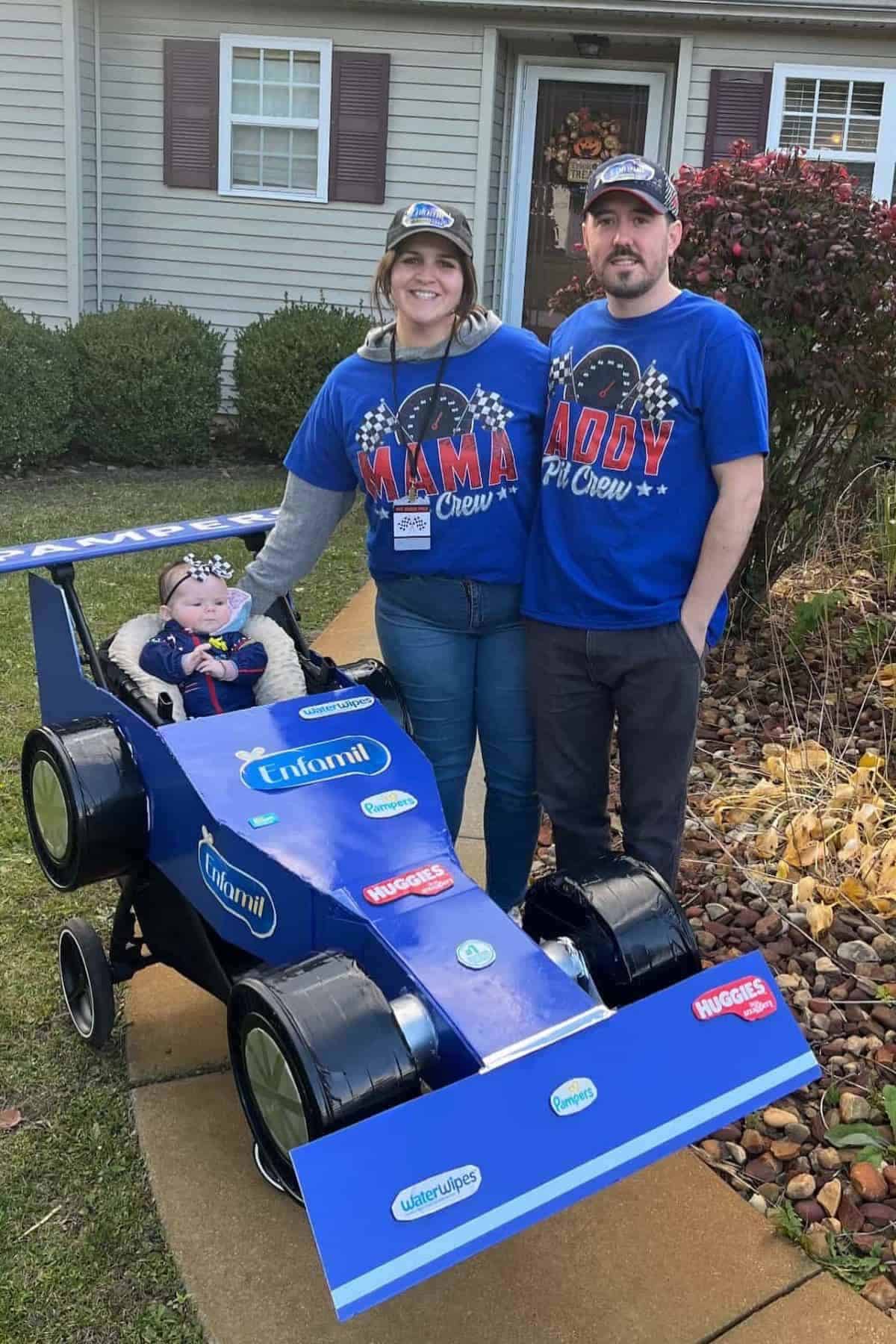 A family stands outside with a stroller decorated as a blue race car. The baby sits in the stroller while the parents, wearing matching pit crew shirts, stand beside it and smile.