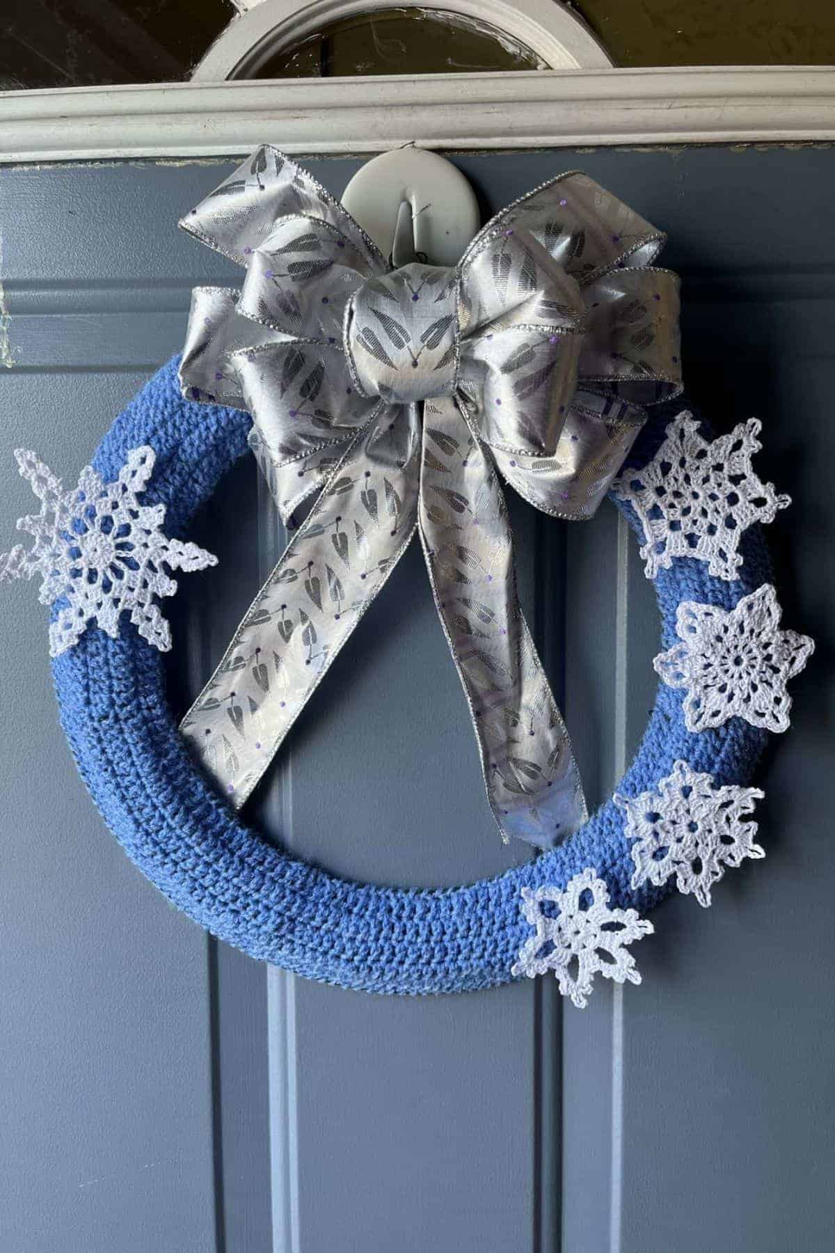 A blue crocheted wreath with white snowflake accents and a large silver bow hangs on a dark door.