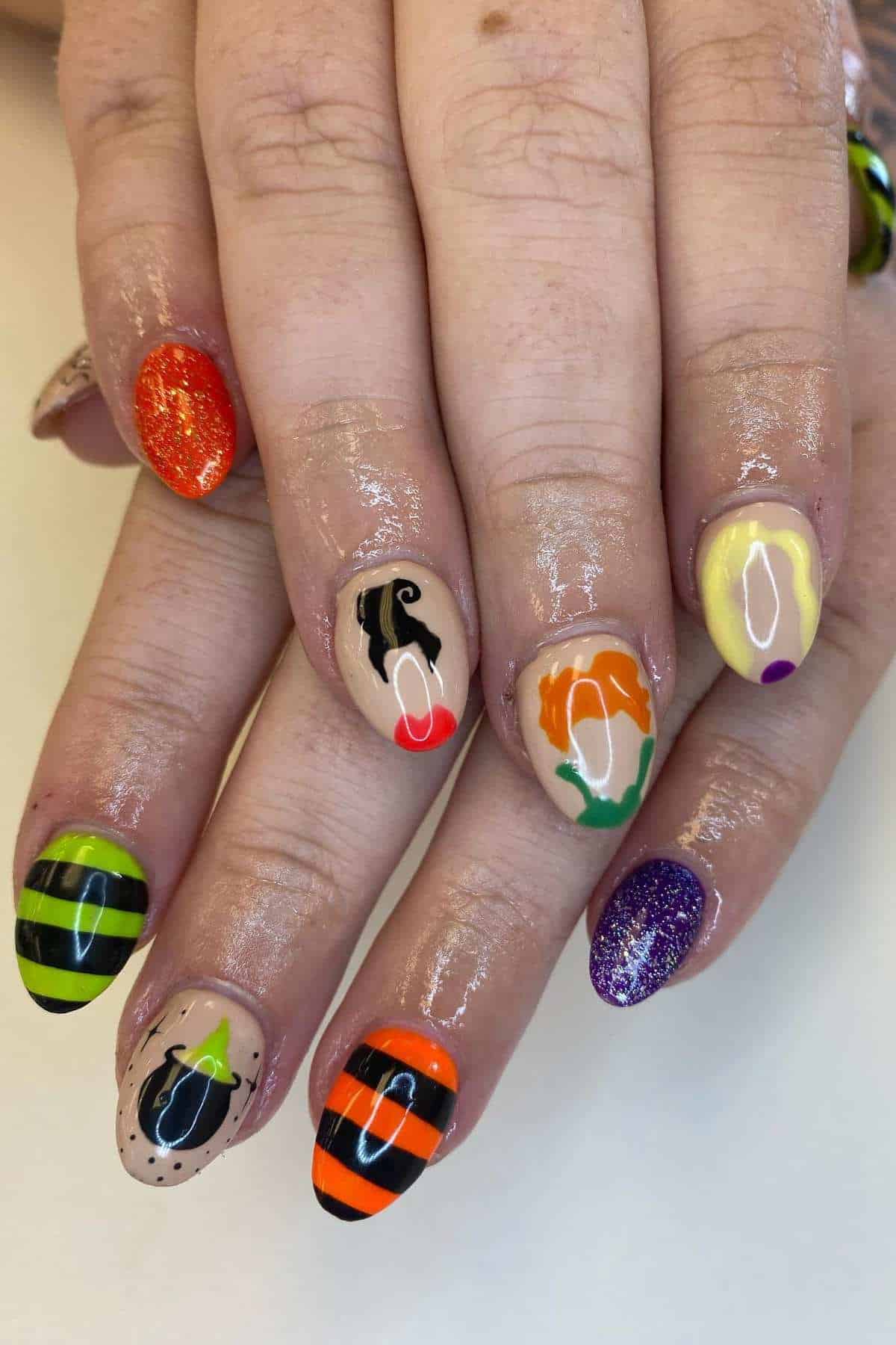 A hand with colorful, Halloween-themed nail art including stripes, glitter, and character designs on each fingernail.