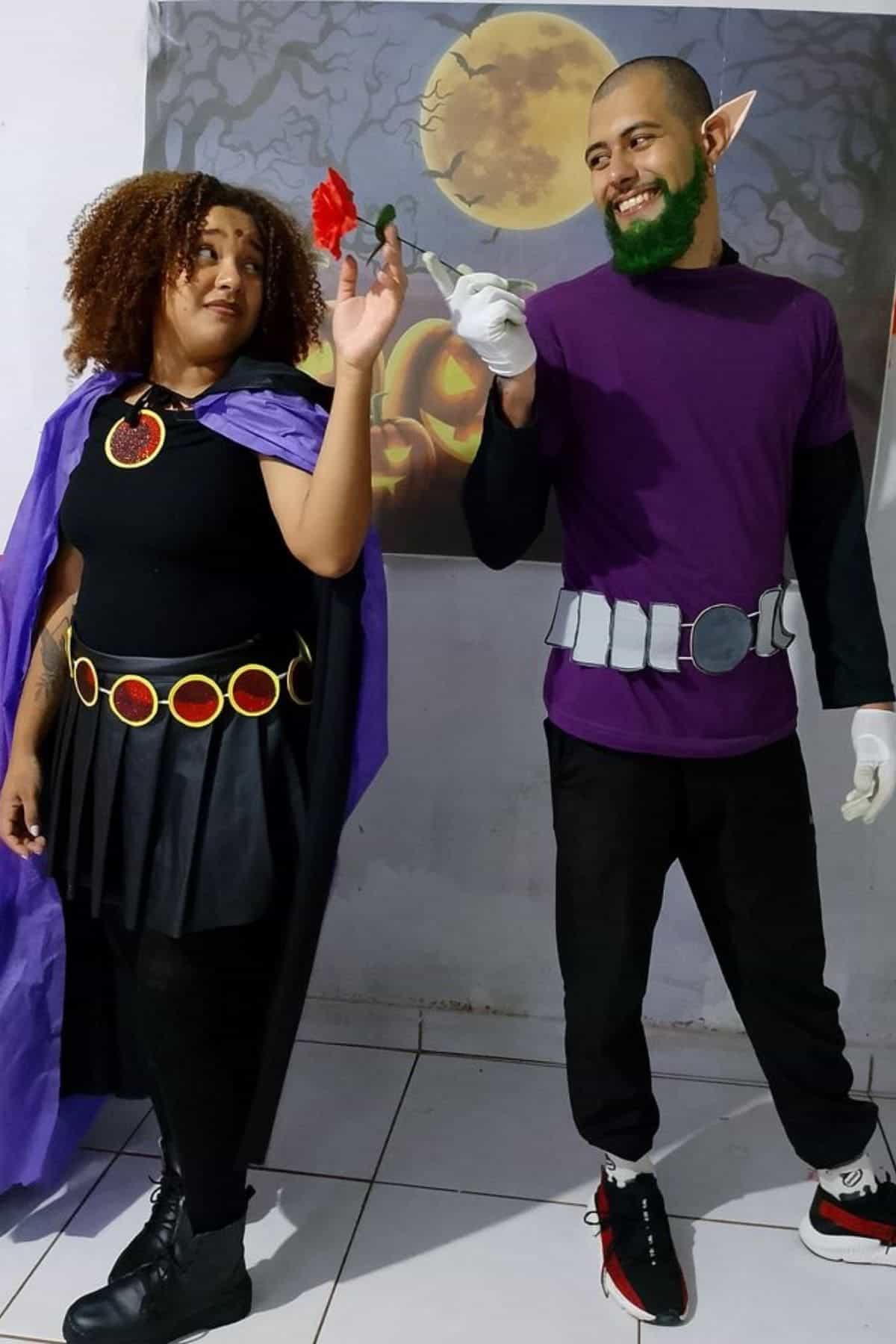 Two people dressed as Raven and Beast Boy from Teen Titans pose together indoors, holding a flower, in front of a wall with a moon-themed painting.