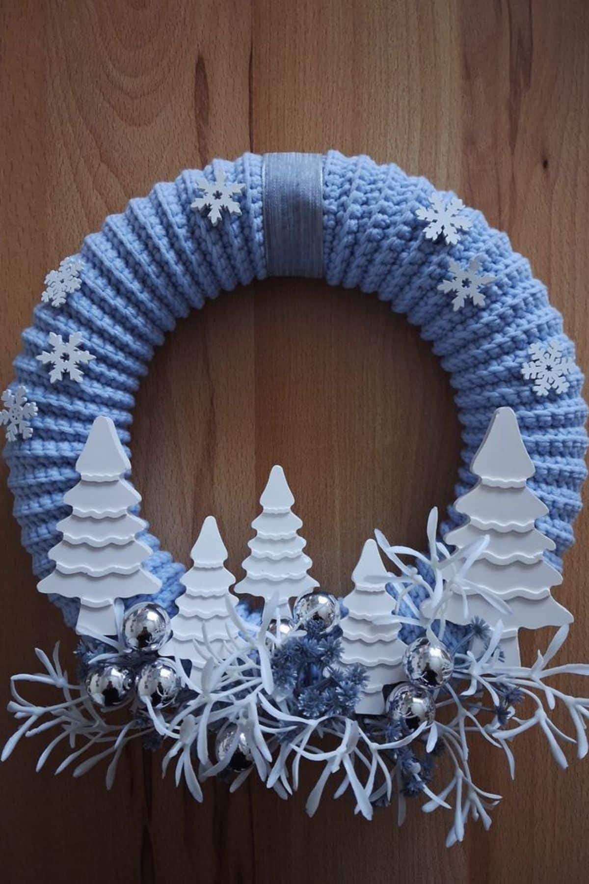 A blue knitted wreath decorated with white artificial trees, silver baubles, snowflakes, and white foliage, displayed on a wooden surface.