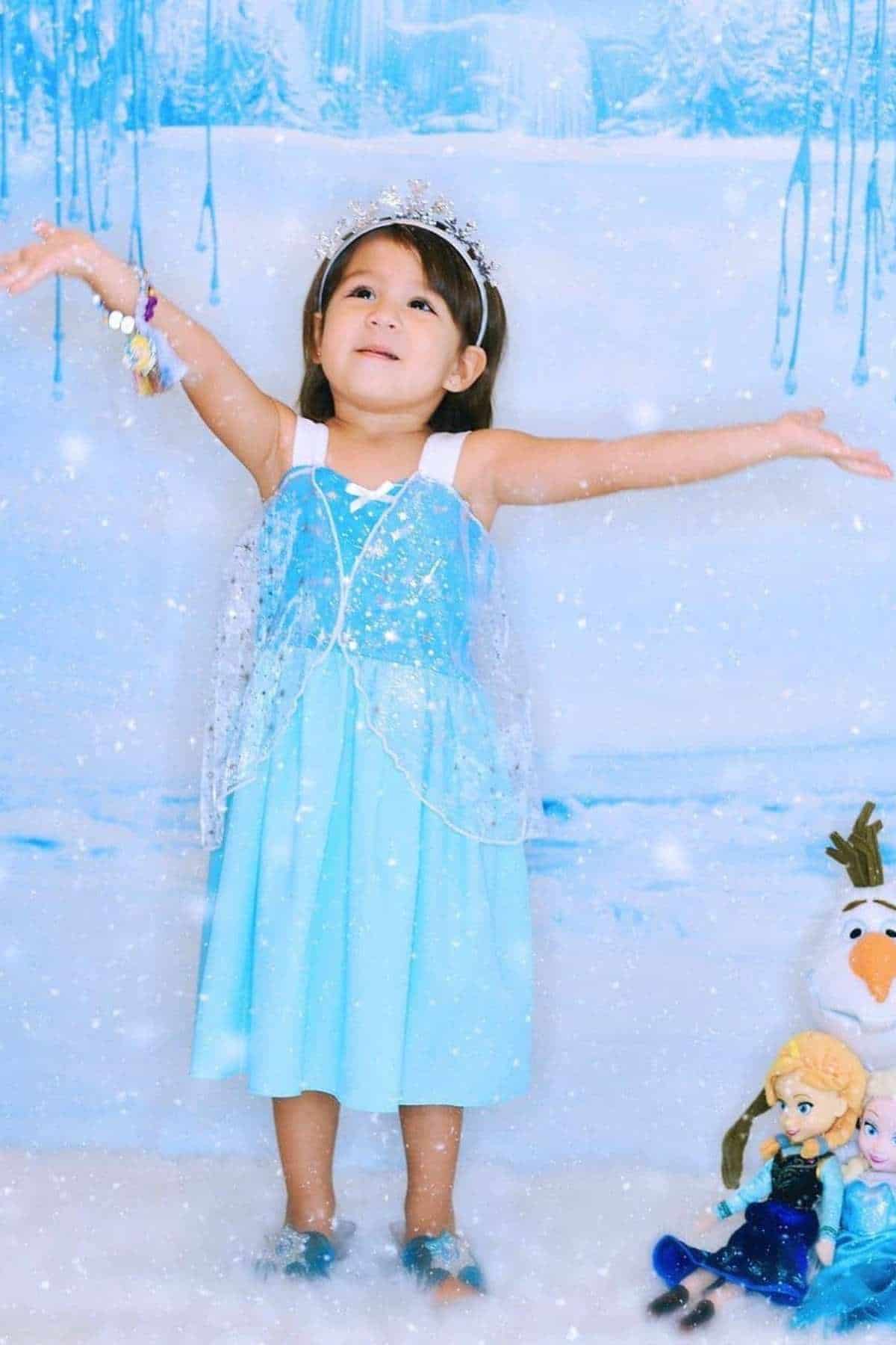 Young girl in a blue princess dress stands with arms outstretched, wearing a crown, surrounded by Frozen-themed dolls and a snowy backdrop.