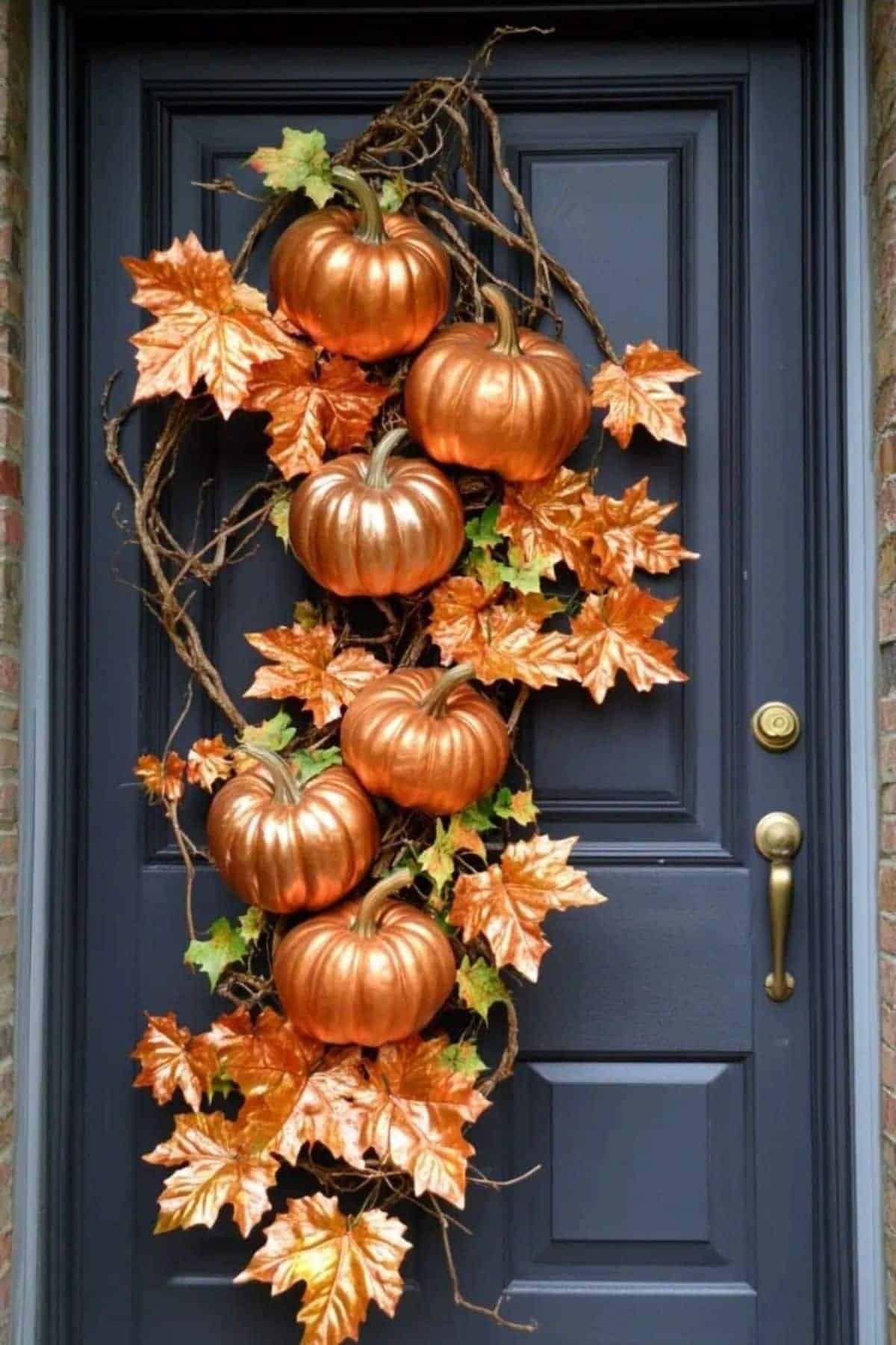 Metallic orange pumpkins and autumn leaves arranged in a vertical garland decorate a dark gray front door with a brass handle.