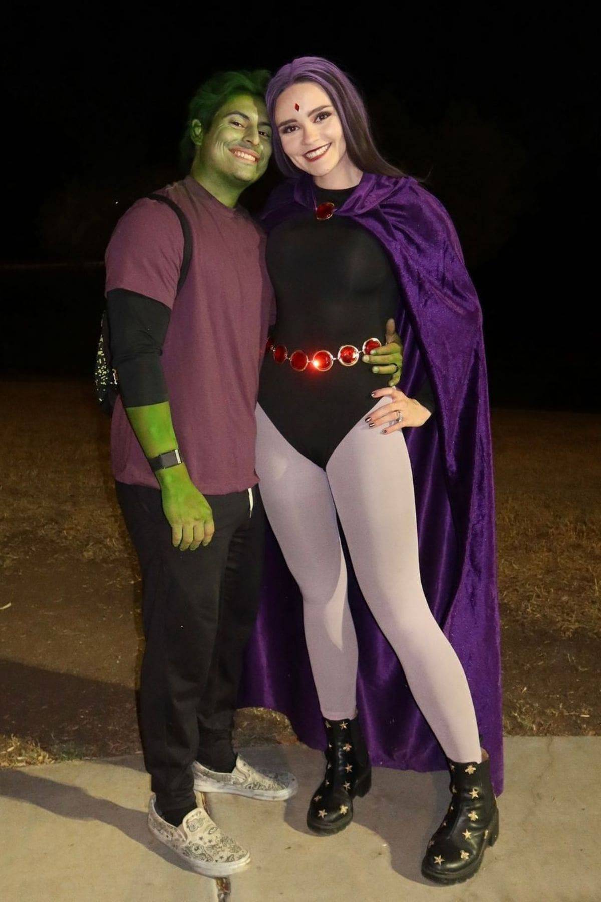 Two people in cosplay stand together. One is dressed as Beast Boy with green face paint; the other is dressed as Raven with a purple cloak and black bodysuit. They are smiling outdoors at night.