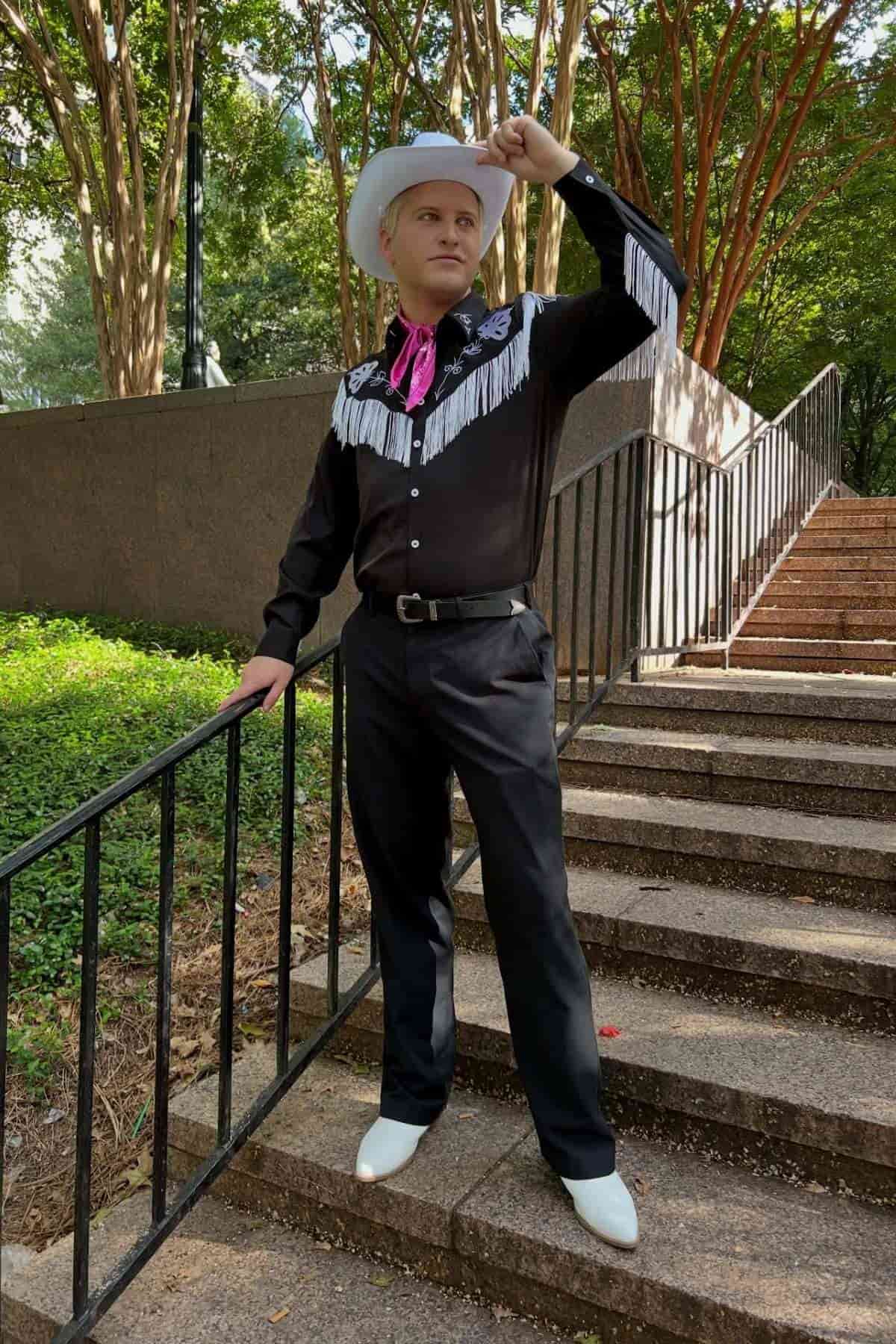 Person in a black western shirt with white fringe, white cowboy hat, and boots stands on outdoor steps, holding the hat brim and posing with one hand on a railing.