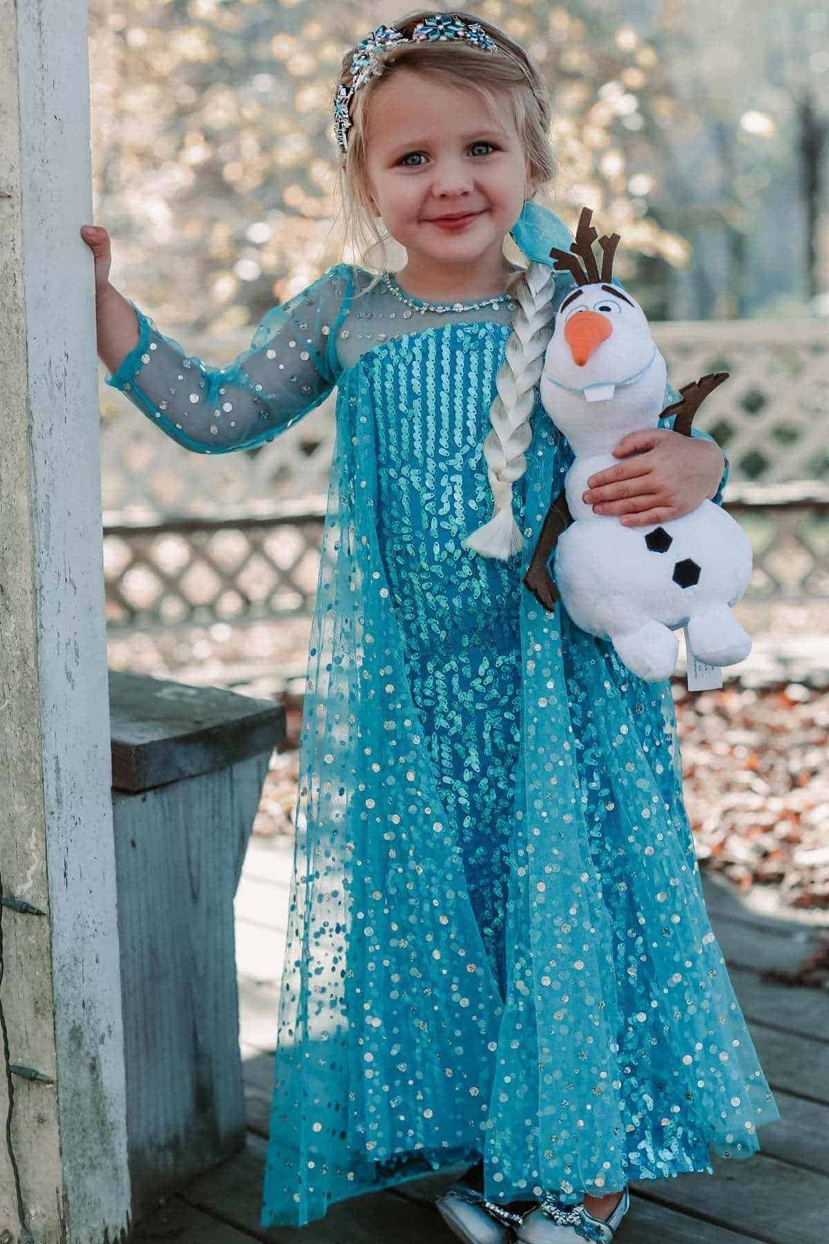 Young girl in a blue sparkly dress and blonde braid stands outdoors, holding a plush snowman toy and smiling at the camera.