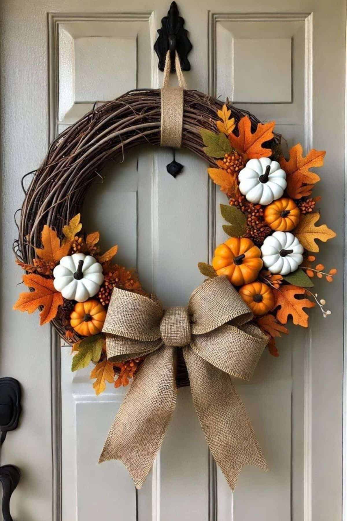 A grapevine wreath with orange and white pumpkins, orange leaves, berries, and a large burlap bow hangs on a beige door.
