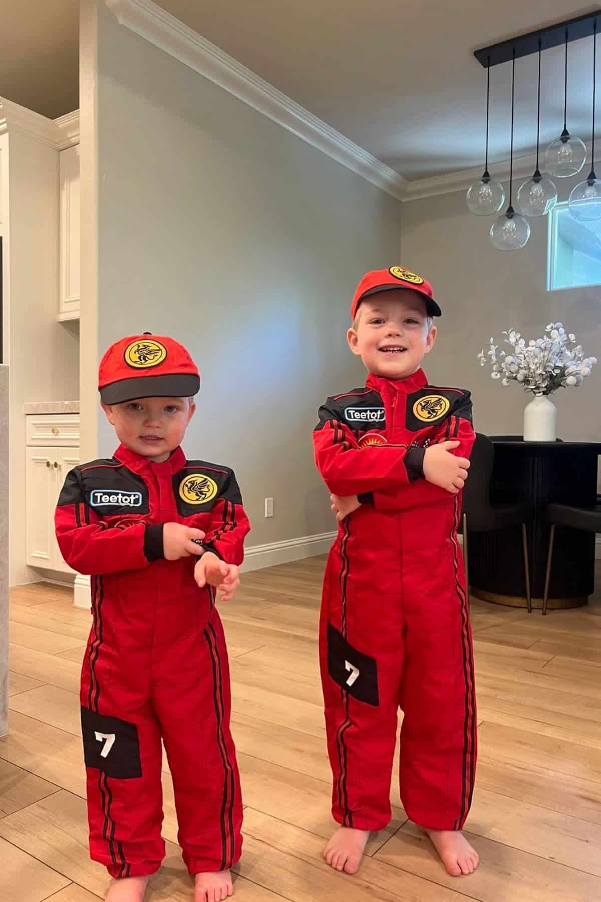 Two young children stand barefoot indoors, wearing matching red racing jumpsuits and hats with patches, in a modern kitchen and dining area.