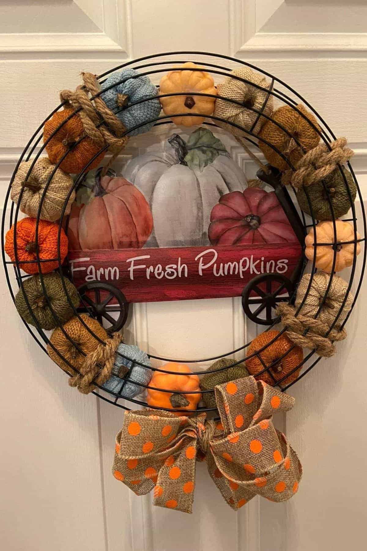 A decorative wreath with fabric pumpkins, a burlap bow, and a sign reading “Farm Fresh Pumpkins” featuring painted pumpkins, hanging on a white door.