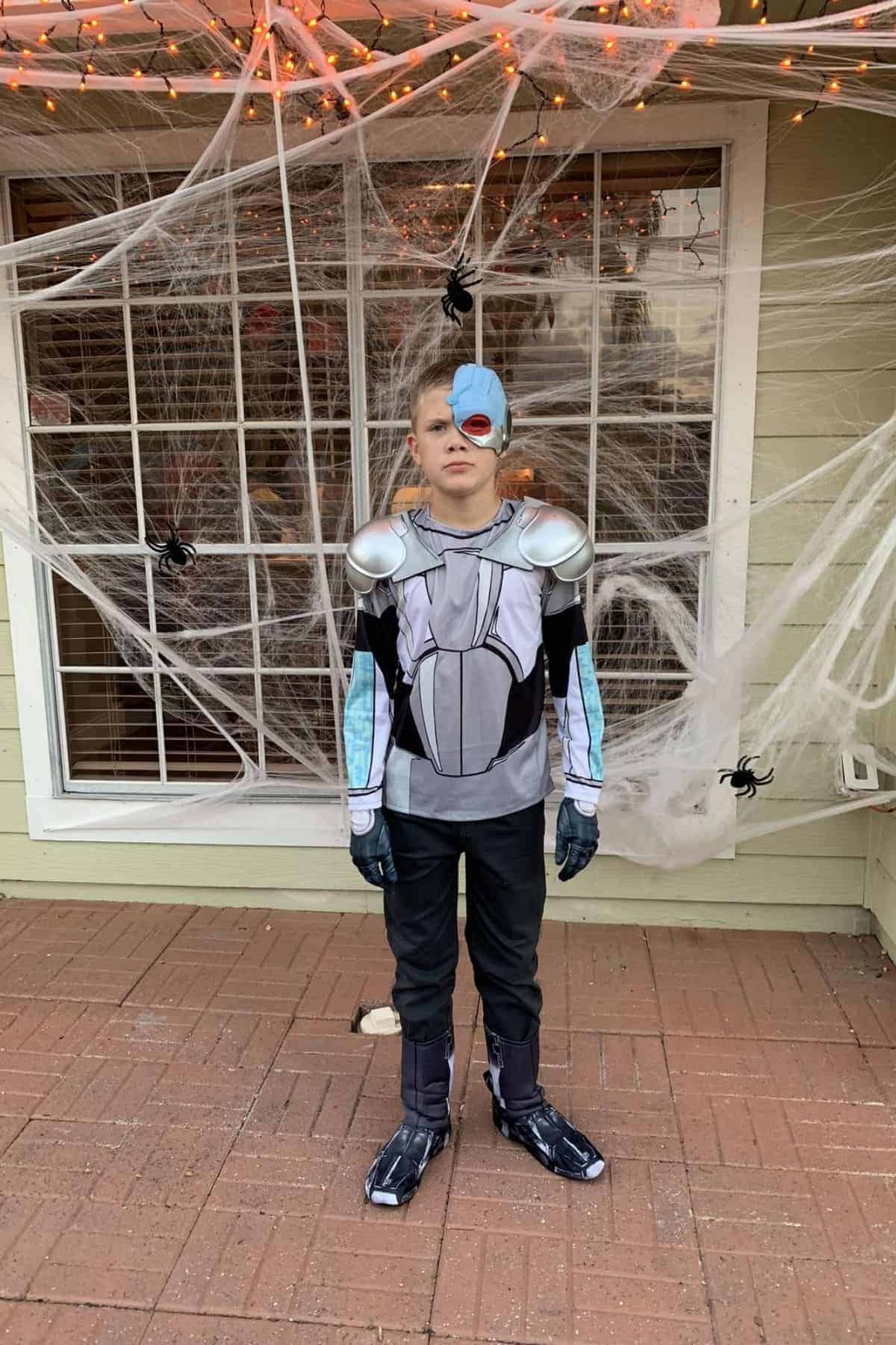 A child in a cyborg costume stands in front of a window covered with fake cobwebs and plastic spiders.