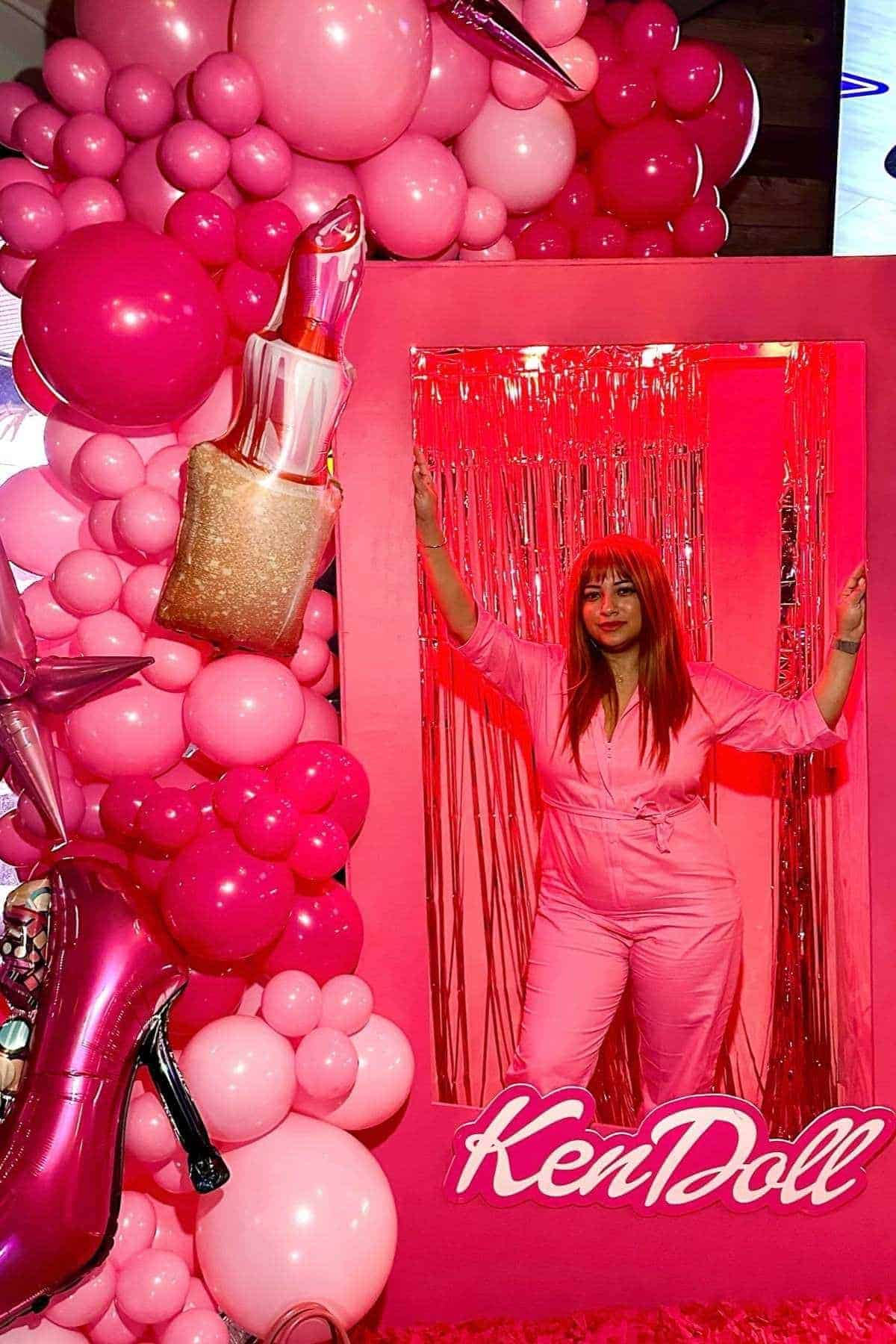 Woman in a pink jumpsuit poses in a box decorated with pink balloons, a lipstick, and high heel props, with “KenDoll” written at the bottom.
