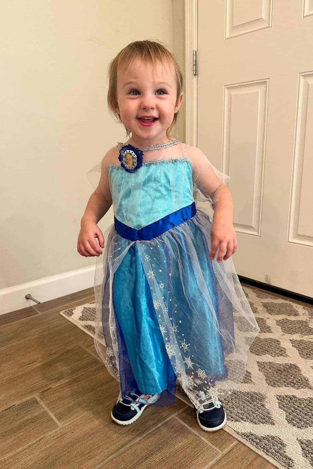 A young child stands indoors, smiling and wearing a blue princess dress with tulle and sneakers, next to a white door on a patterned rug and tiled floor.