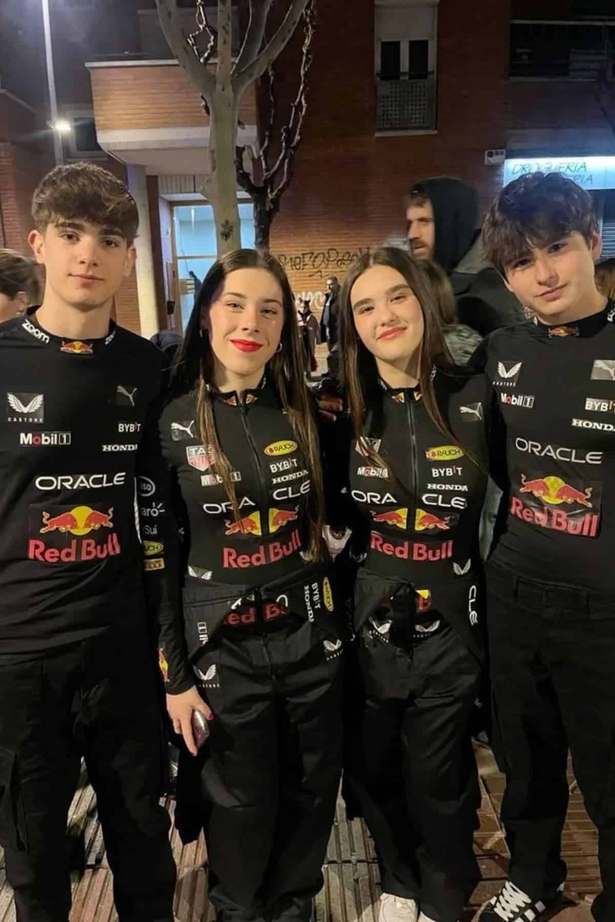 Four young people stand outdoors at night, wearing matching black Red Bull Racing uniforms, smiling for the camera. Buildings and other people are visible in the background.