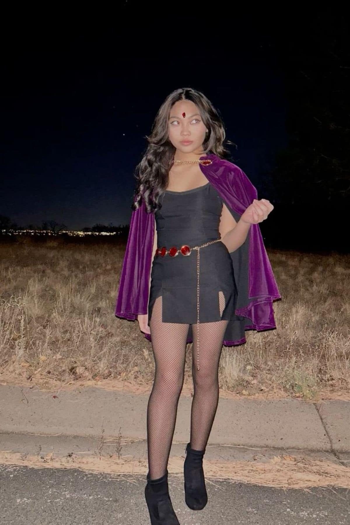 Person wearing a dark dress, purple cape, belt with red gems, fishnet tights, and boots stands on a roadside at night with a field in the background.