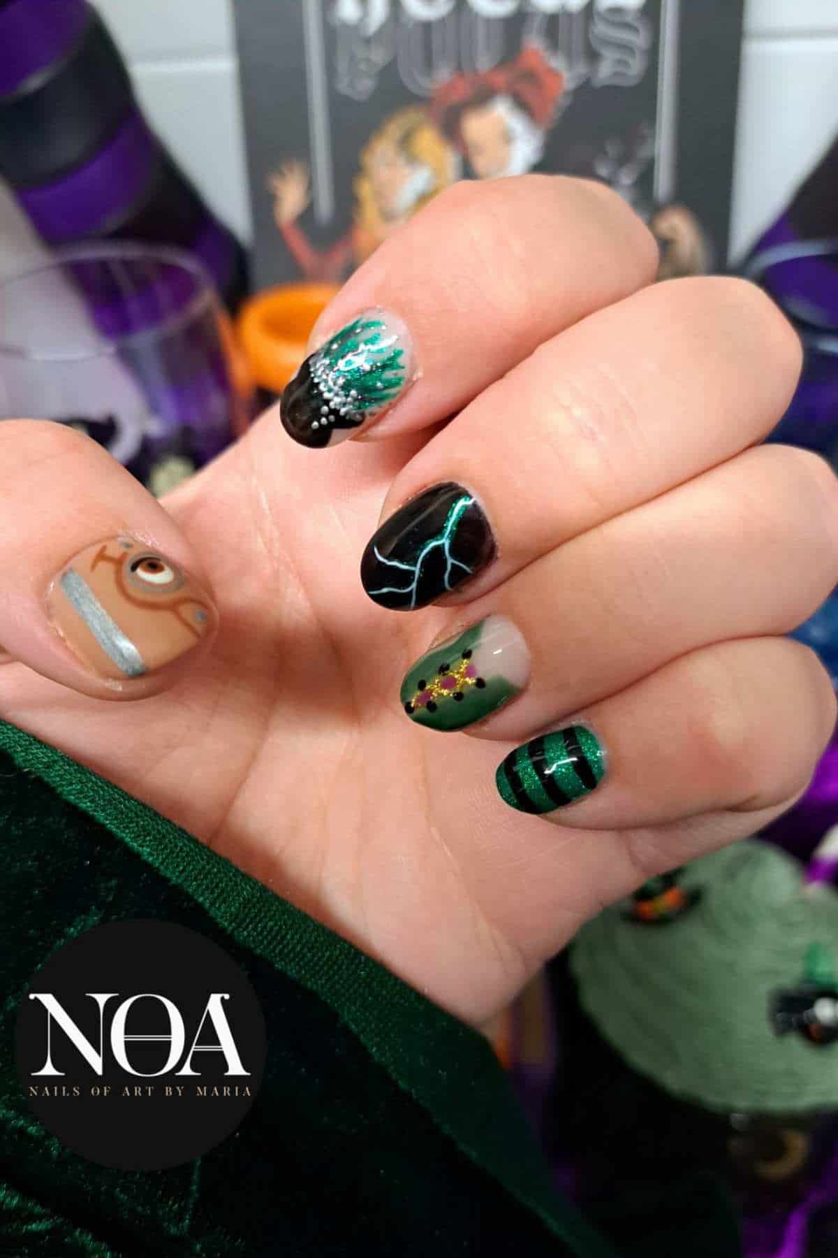 A hand with Halloween-themed nail art, each nail featuring different designs including a green dress, lightning, an eye, and black and green stripes. Background includes themed decorations.