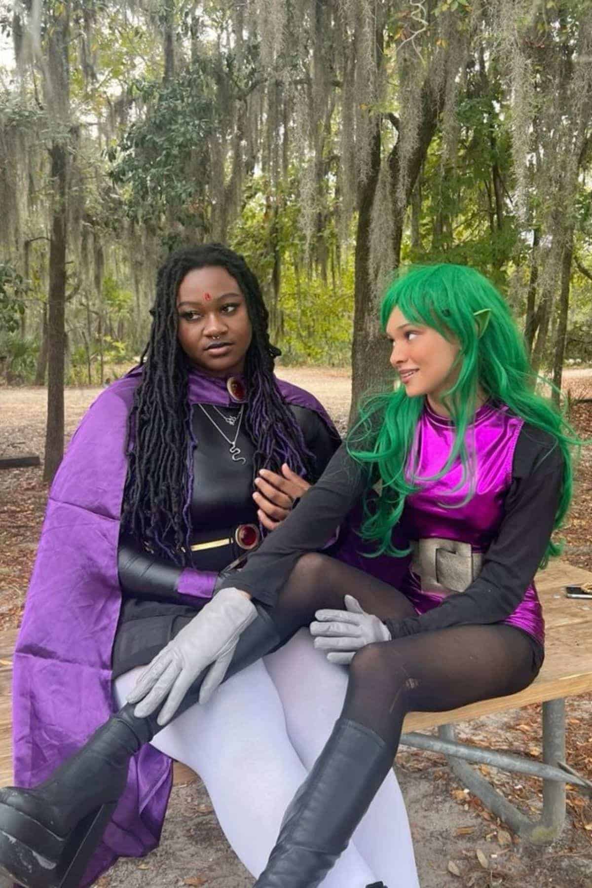 Two people in colorful cosplay costumes sit on a wooden bench outdoors, surrounded by trees and hanging moss, engaging in conversation.