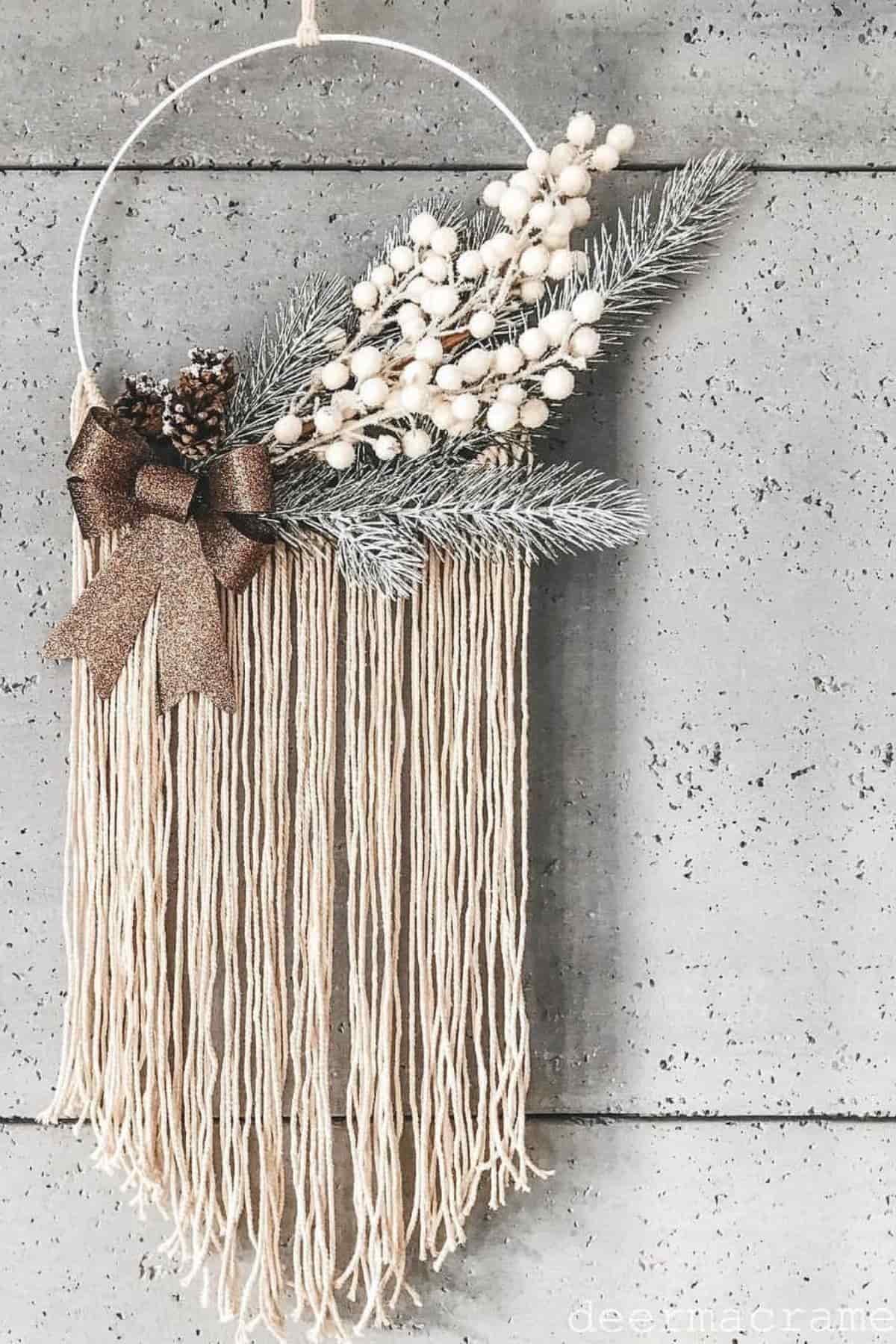 A handmade wall wreath with cream yarn strands, pine needles, white berries, pinecones, and a brown ribbon bow, hanging on a gray wooden wall.