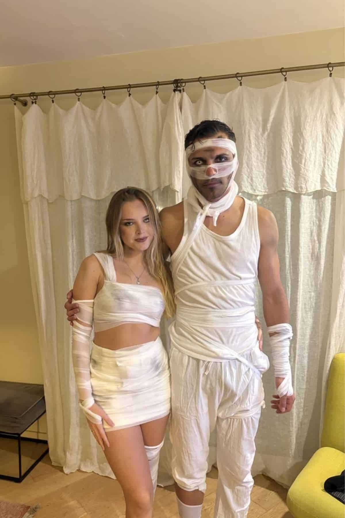 Two people dressed in white, mummy-inspired costumes stand together indoors in front of light curtains, posing for a photo.