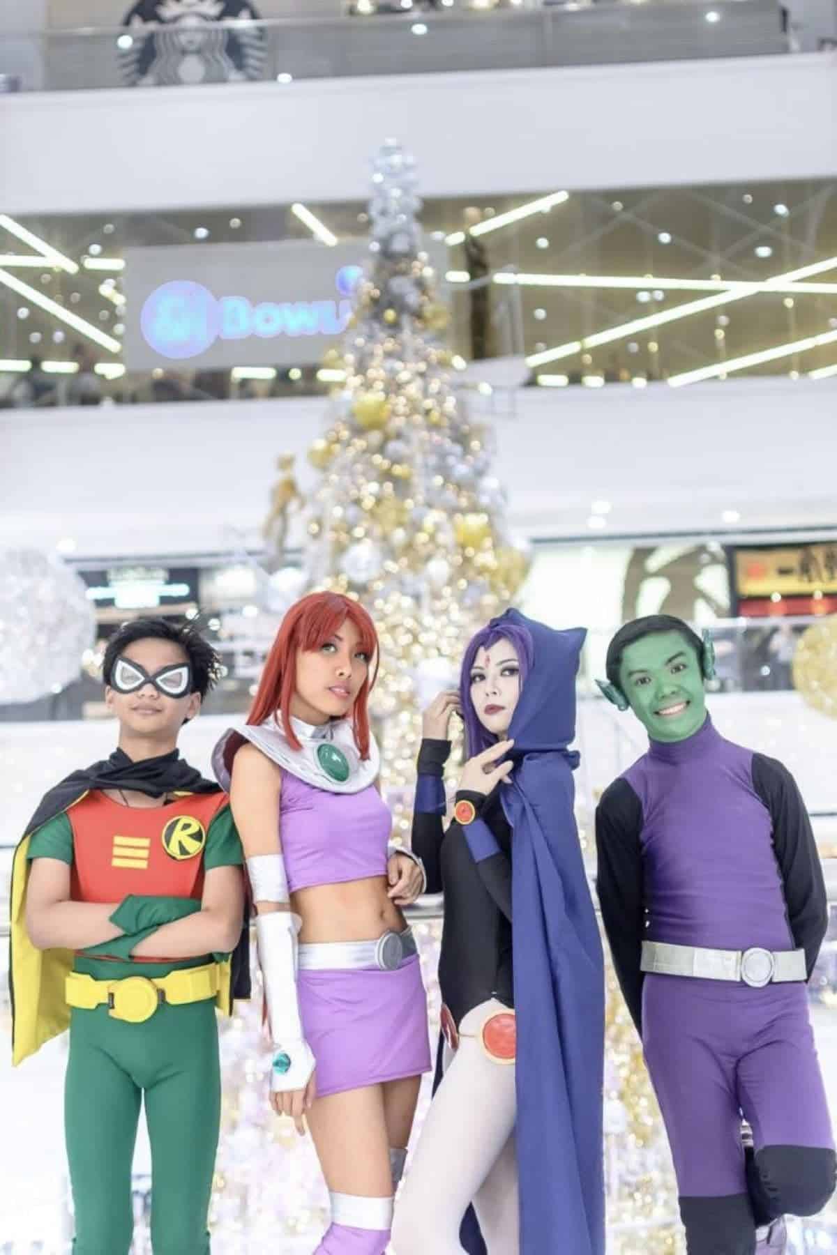 Four people in Teen Titans costumes pose in front of a white and gold Christmas tree in a modern indoor mall with festive decorations.