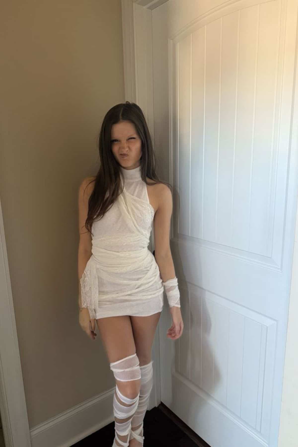 A person in a white, mummy-inspired costume stands indoors by a white door, making a scrunched facial expression.