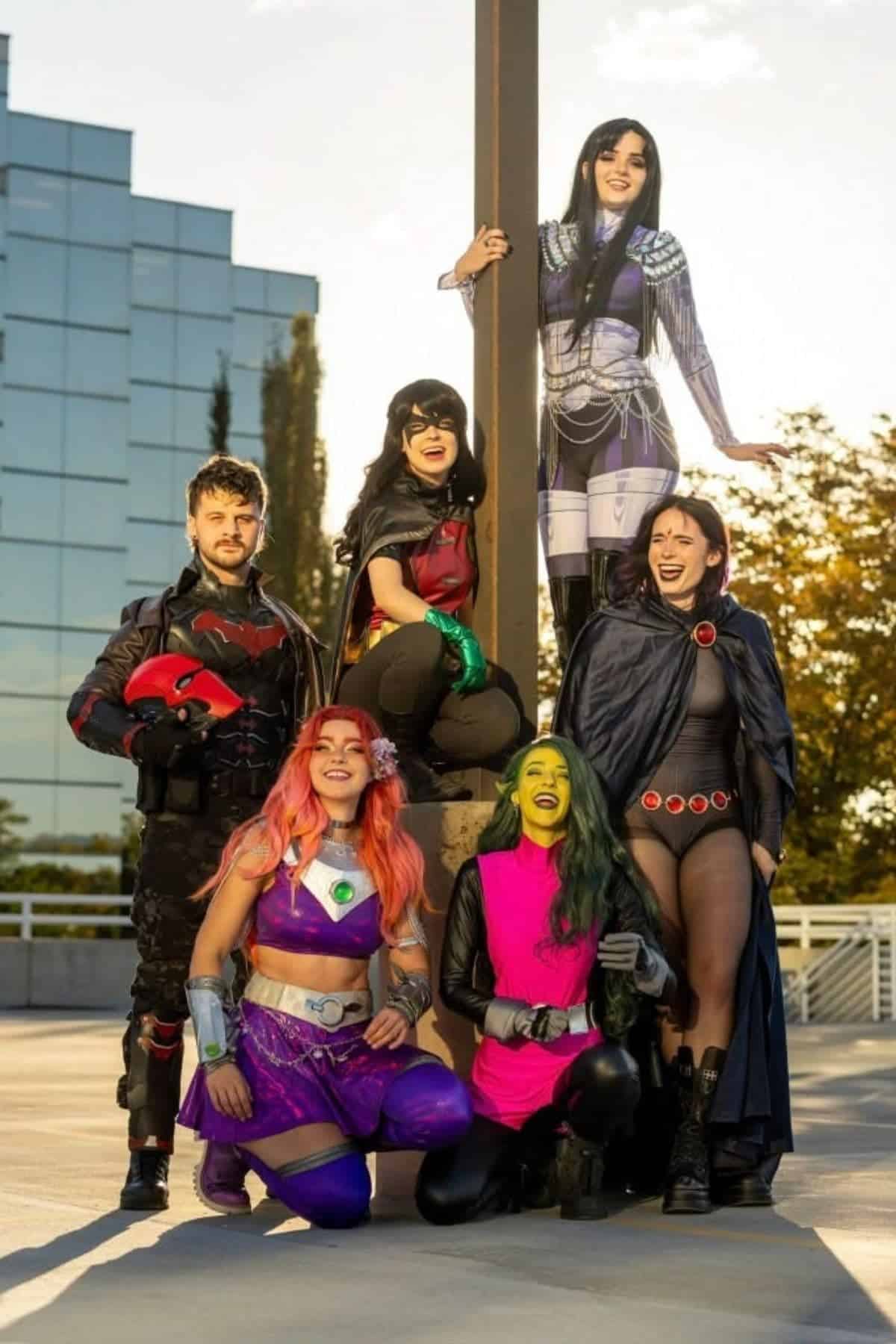 Six people in detailed cosplay costumes pose outdoors near a lamp post, with a modern glass building and trees in the background.