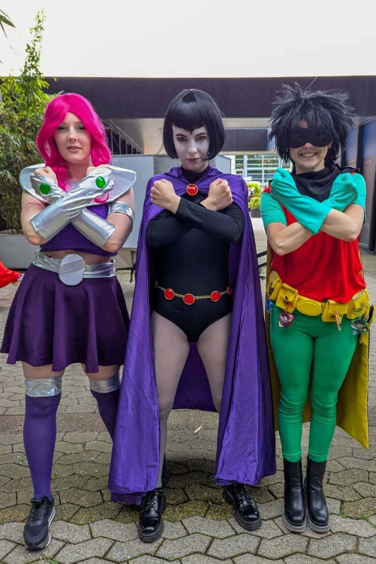 Three people in Teen Titans costumes pose outdoors: one as Starfire, one as Raven with arms crossed, and one as Robin with arms behind their back.