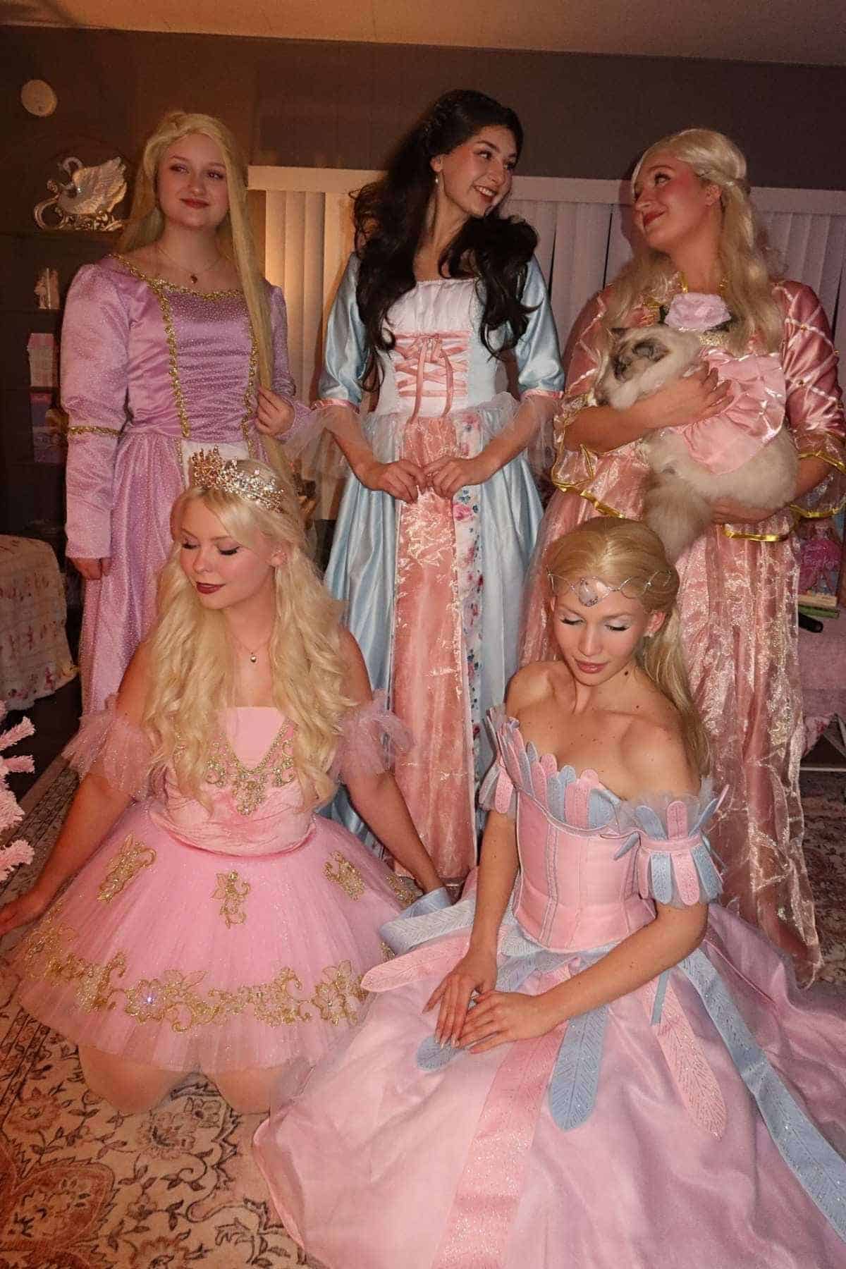 Five women dressed in elaborate princess costumes pose indoors; one holds a cat while others stand or sit, all wearing pastel-colored gowns.