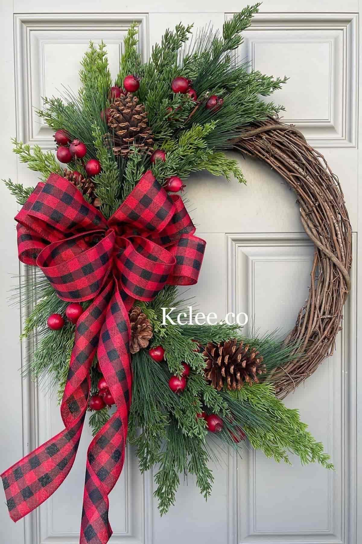 A wreath with pine branches, red berries, pinecones, and a red plaid bow hangs on a white door.
