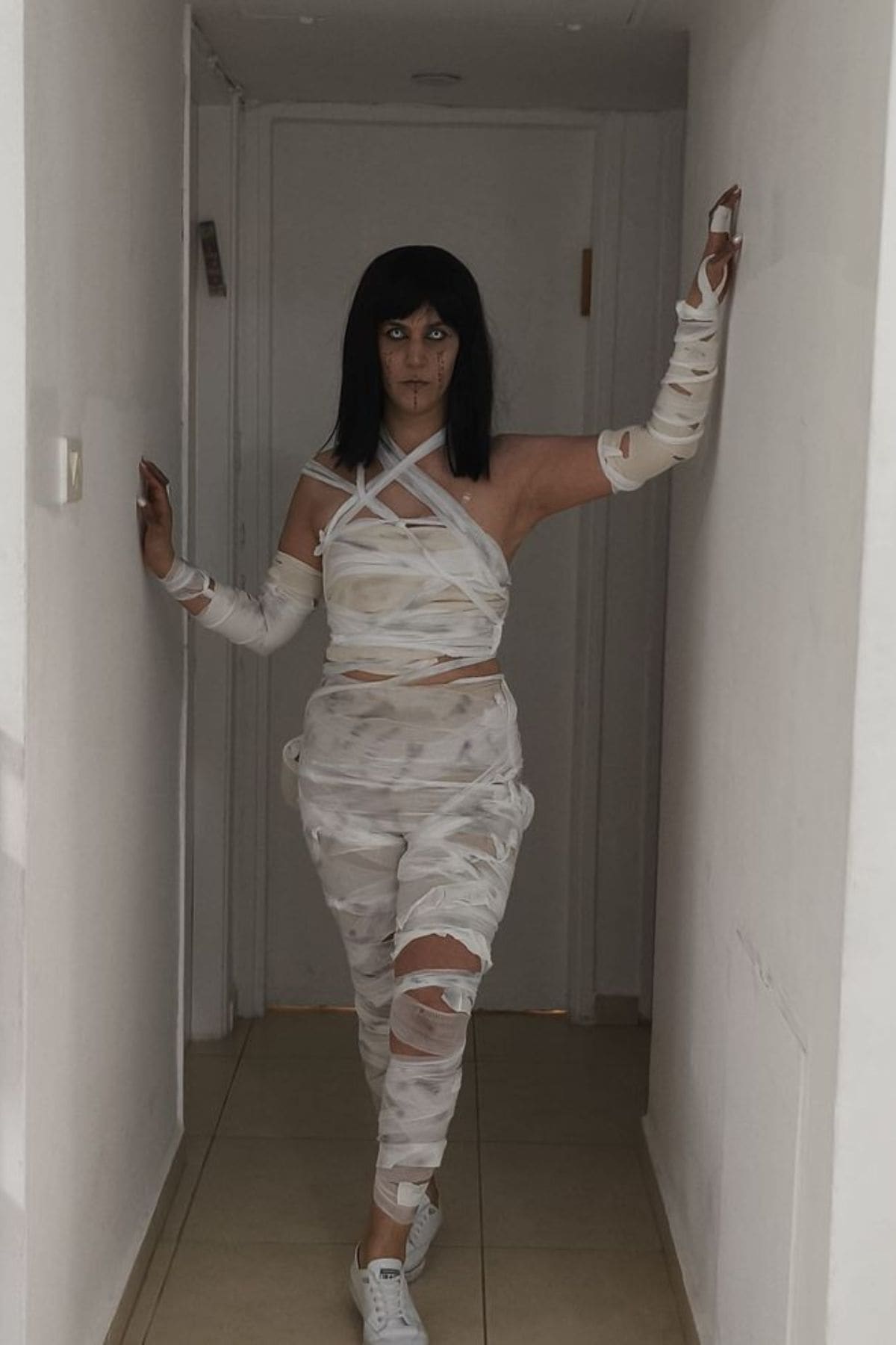 Person in a hallway wearing a costume made of white bandages, standing with arms outstretched, looking at the camera.