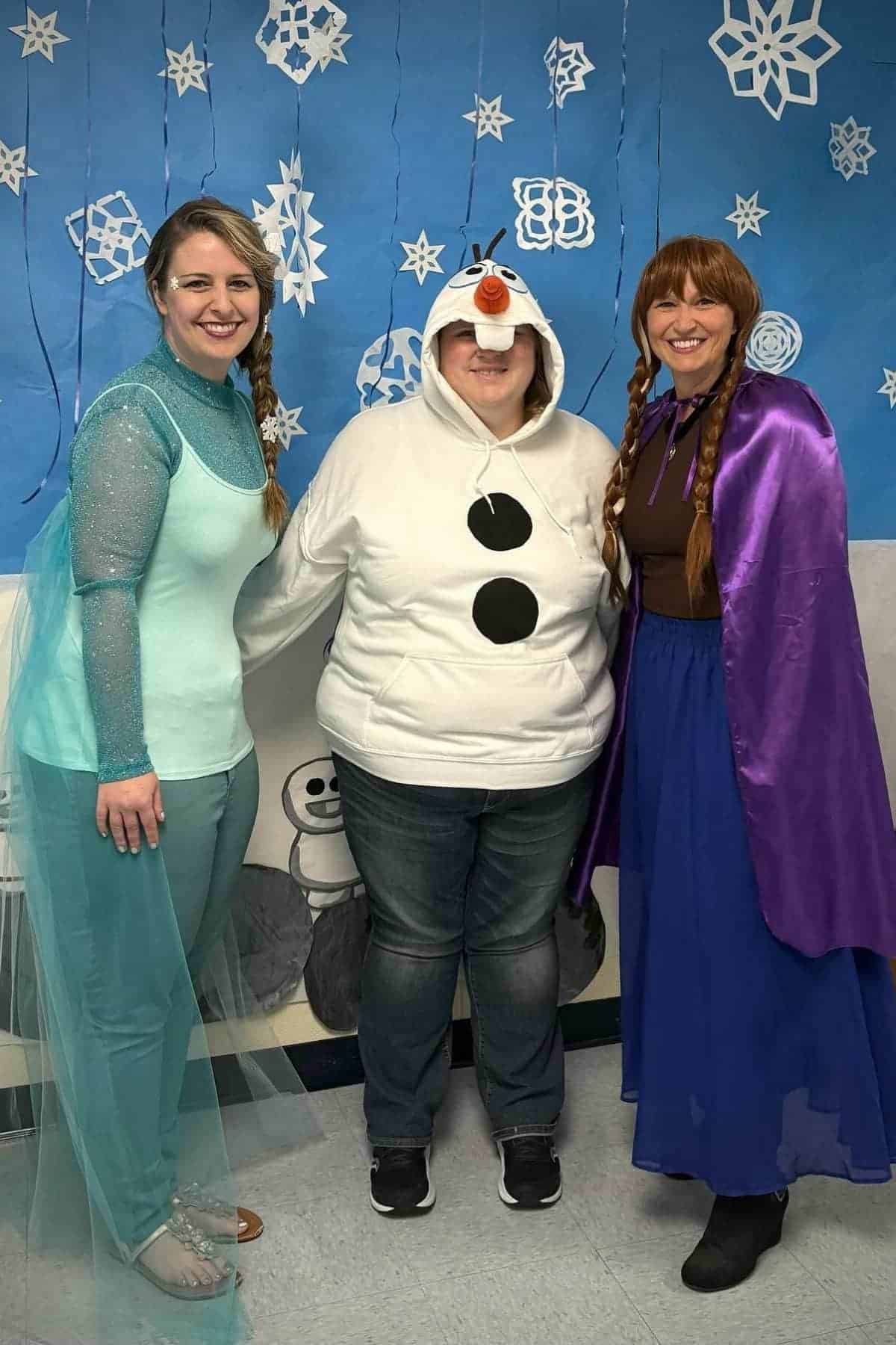 Three adults in costumes pose in front of a snowflake-themed backdrop; two as Frozen characters Elsa and Anna, one in a white Olaf hoodie.