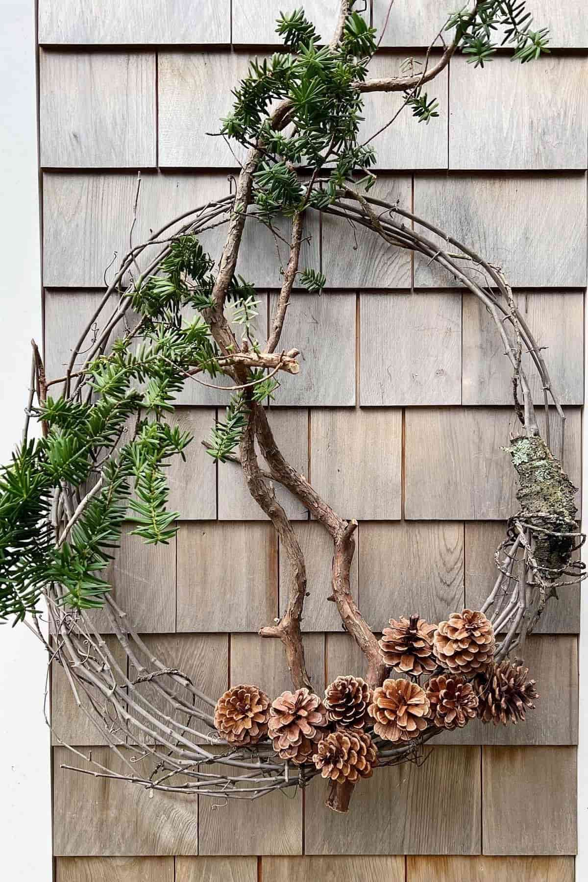 A wreath made of twigs, branches, and pinecones hangs on a wood shingle wall, with sprigs of greenery and a patch of lichen attached.