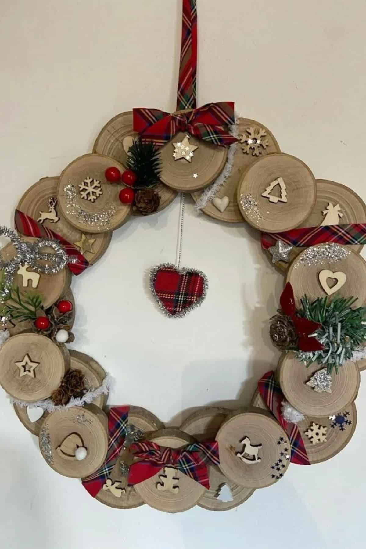 Wooden wreath decorated with plaid bows, small pine branches, red berries, silver garland, and wooden holiday shapes, hanging on a wall with a plaid heart ornament in the center.