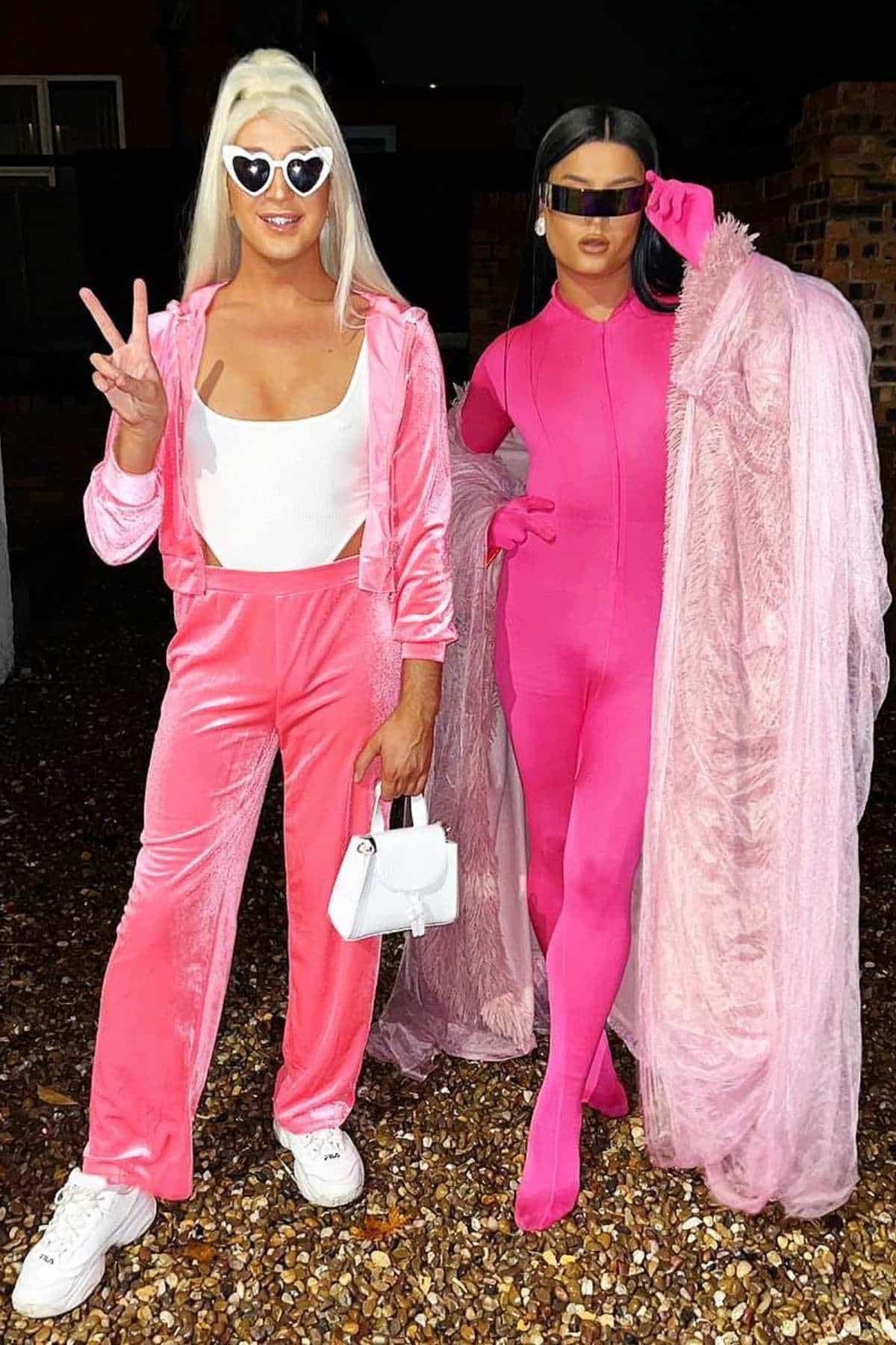 Two people pose outdoors wearing bright pink outfits; one in a tracksuit with heart sunglasses, the other in a fitted bodysuit with a long sheer robe and futuristic sunglasses.