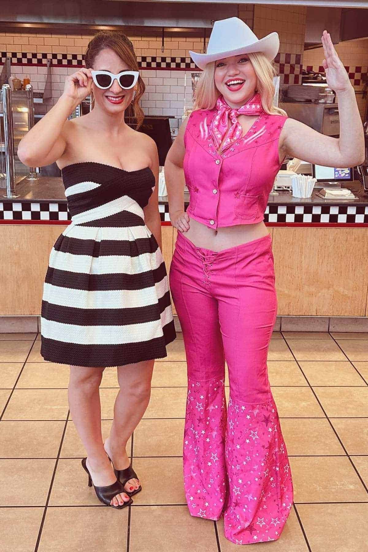 Two women stand indoors. One wears a black-and-white strapless dress and sunglasses; the other wears a pink cowboy outfit with a vest, flared pants, scarf, and hat, smiling and waving.