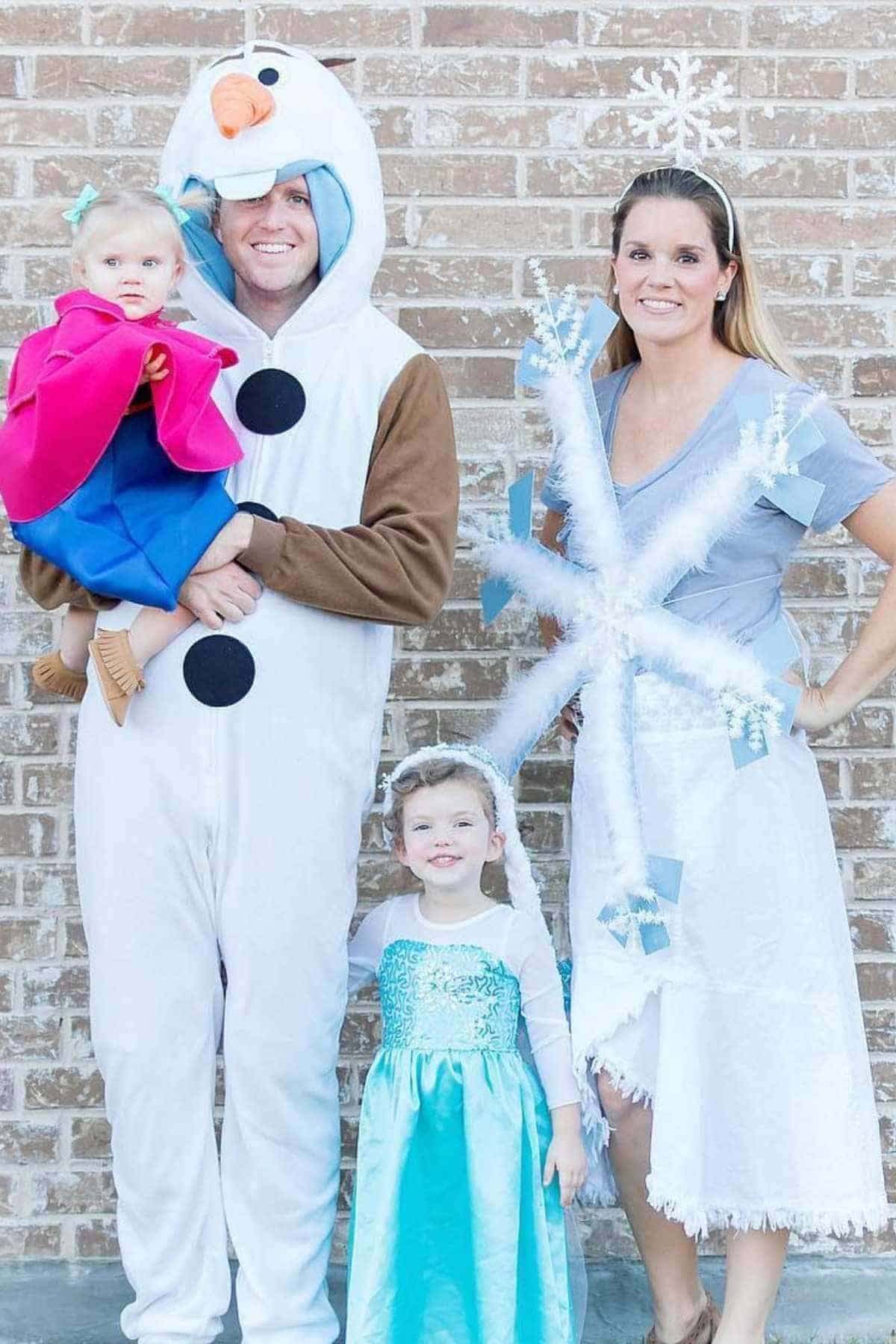 Four people wearing Frozen-themed costumes pose in front of a brick wall; one adult is Olaf, another is a snowflake, and two children are dressed as Elsa and Anna.