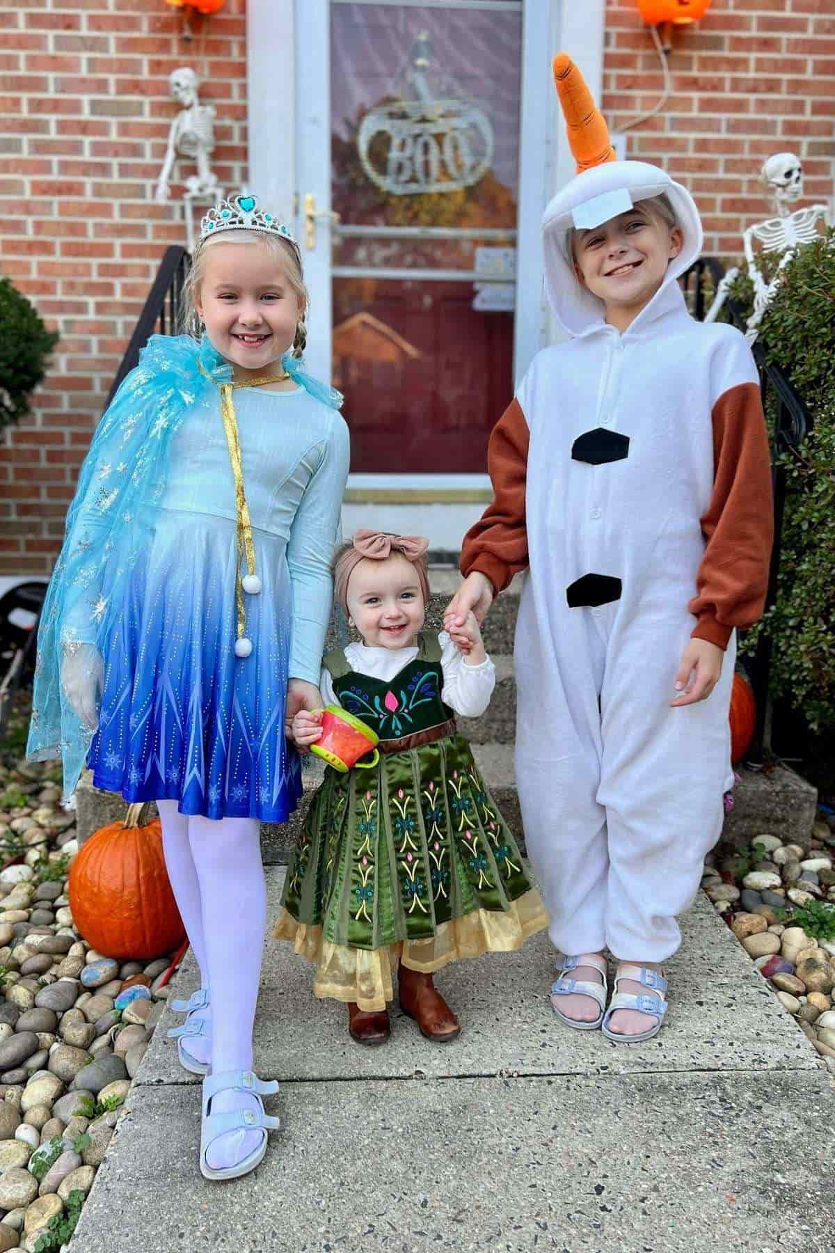 Three children in Frozen-themed costumes stand outside a house decorated for Halloween, with pumpkins and skeletons by the door.