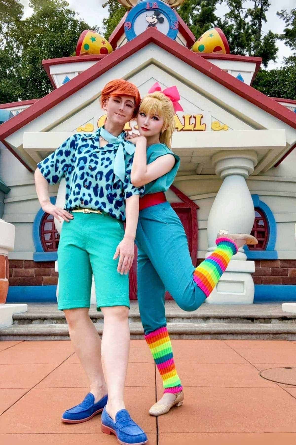 Two people in colorful costumes pose in front of a cartoon-style building; one wears teal shorts and a blue shirt, and the other wears a teal jumpsuit with rainbow leg warmers.